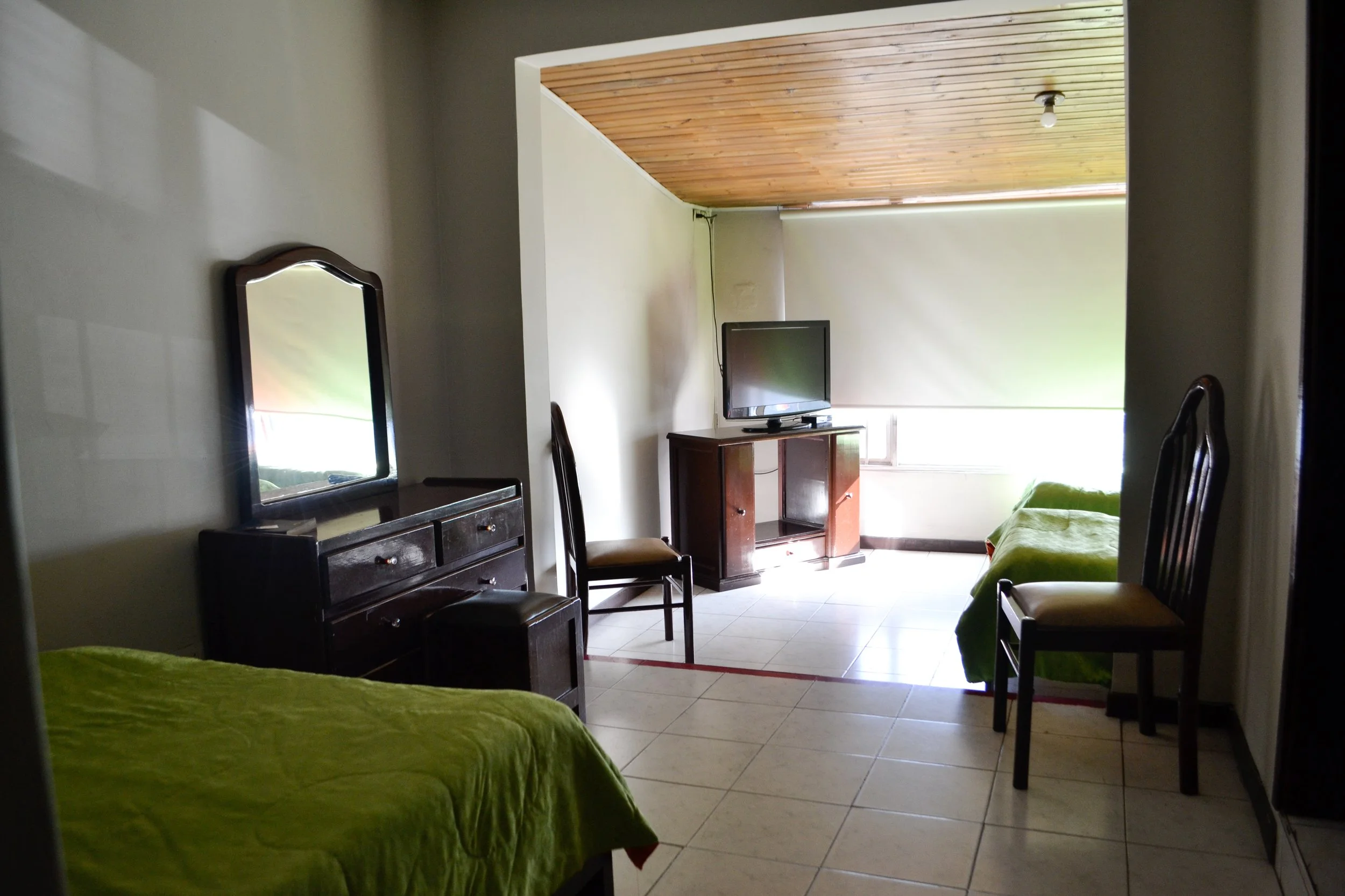 Habitación con dos camas, un tocador con espejo, sillones, televisión y ventana con cortina beige, techo de madera, piso de baldosas claras.