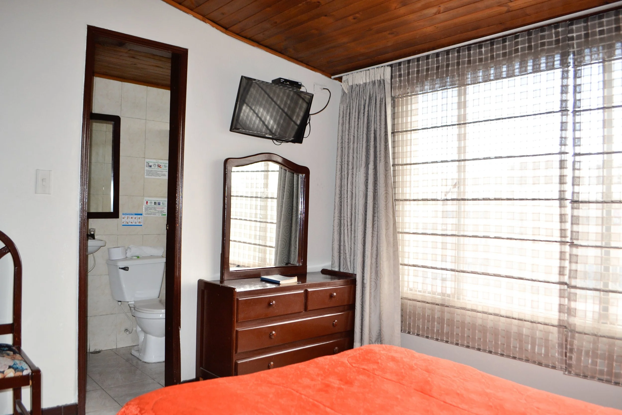 Vista de un habitación con ventana grande con cortinas, una cama con cubrecama naranja, un espejo, un mueble con cajones y un televisor en la pared