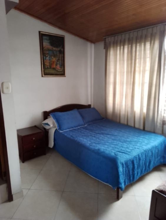 Cama con sábanas azules junto a una mesita de noche en una habitación con cortinas beige y pintura en la pared