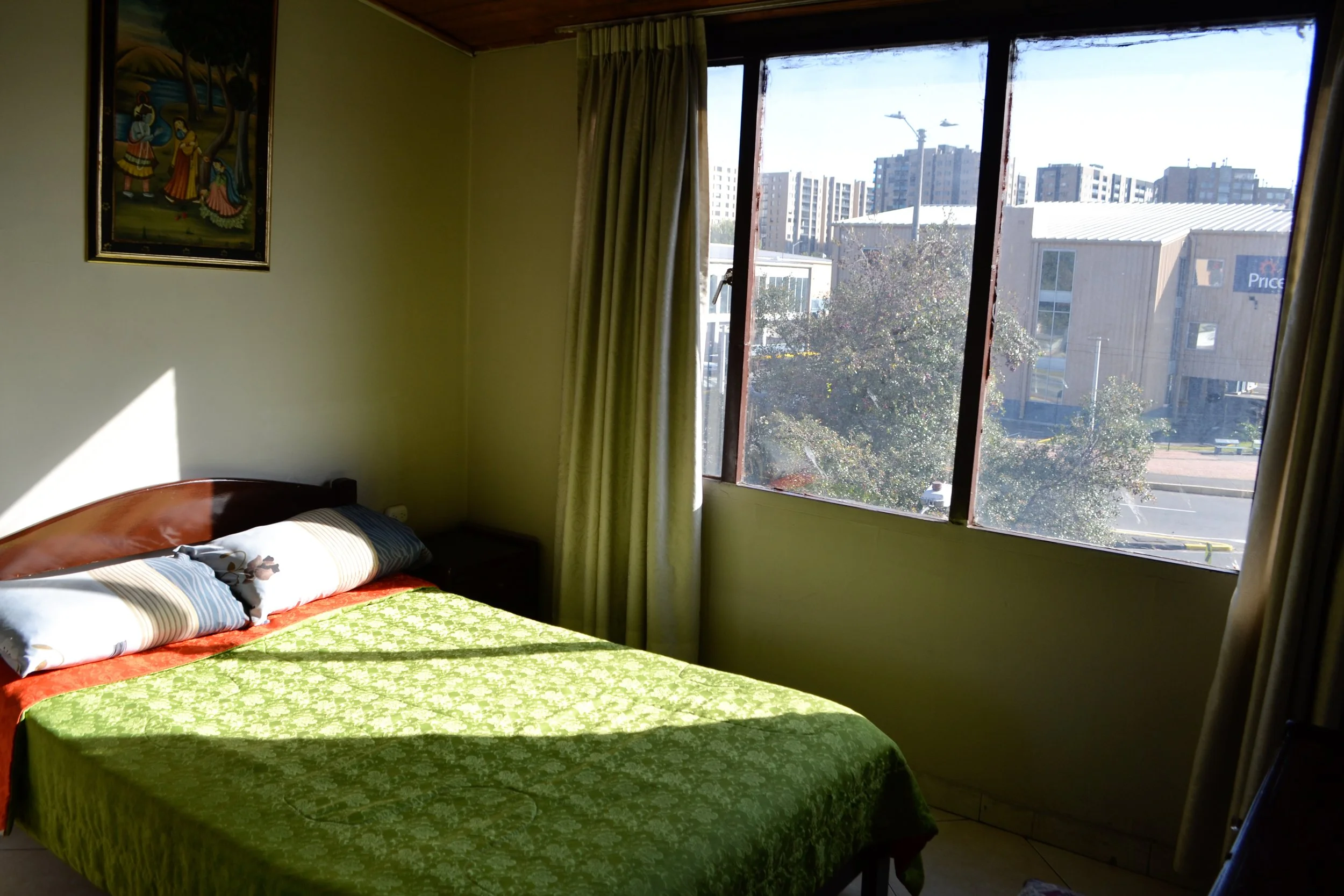 Habitación con cama, edredón verde, dos almohadas, ventana grande con vista a edificios y árboles, cortinas beige, cuadro en la pared.