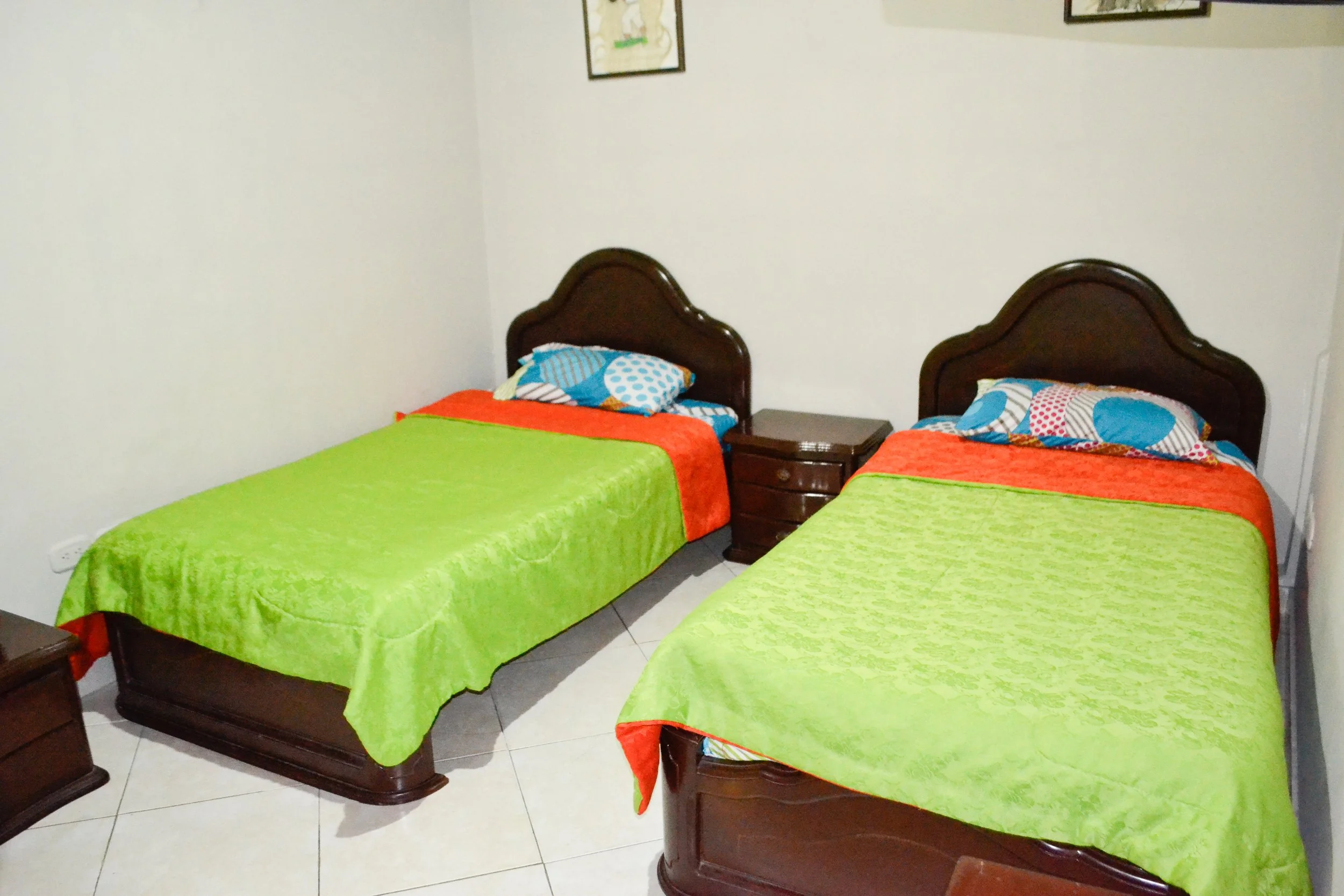 Dos camas individuales con cabeceros de madera, ropa de cama verde y mantas naranjas, en una habitación con paredes blancas y cuadros colgados.