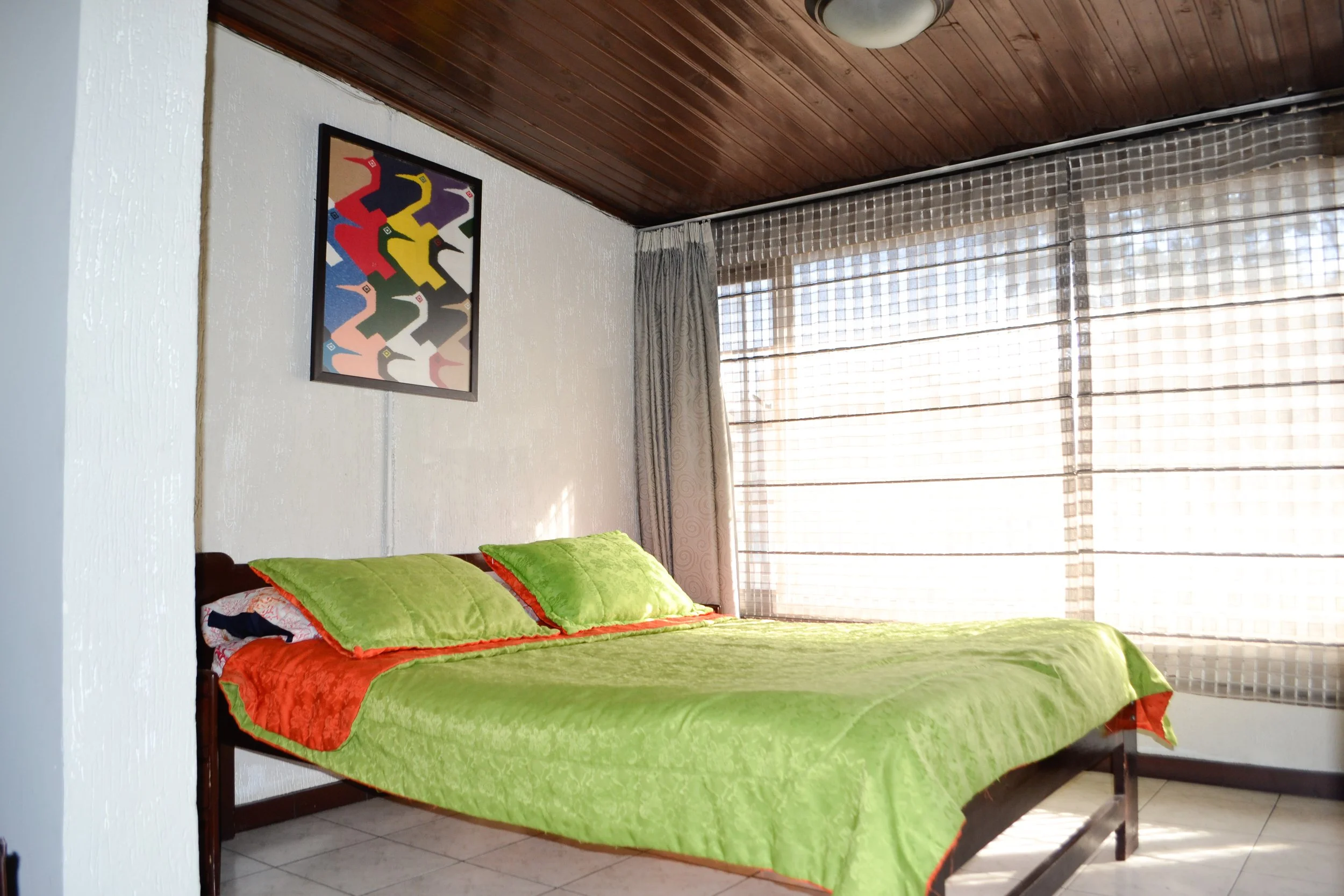 Habitación con cama de madera, sábanas verdes y almohadas verdes, cortinas blancas y ventanas con toldos de listones y cortinas. Obra de arte abstracto en la pared.