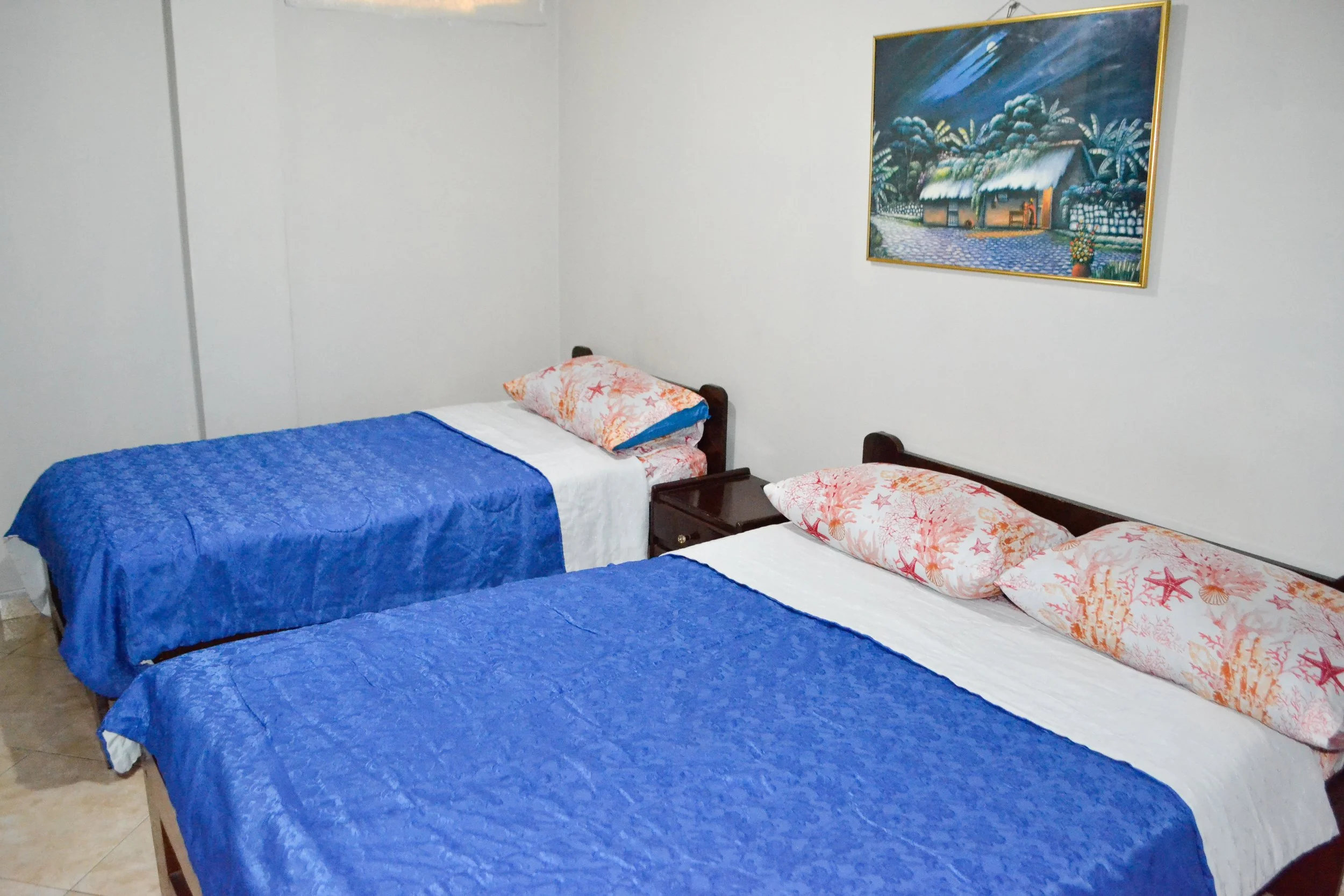 Habitación con dos camas con sábanas azules y almohadas con fundas de colores con conchas y estrellas de mar. Hay una pintura en la pared que muestra una casa en un paisaje tropical con palmeras y un cielo nocturno.