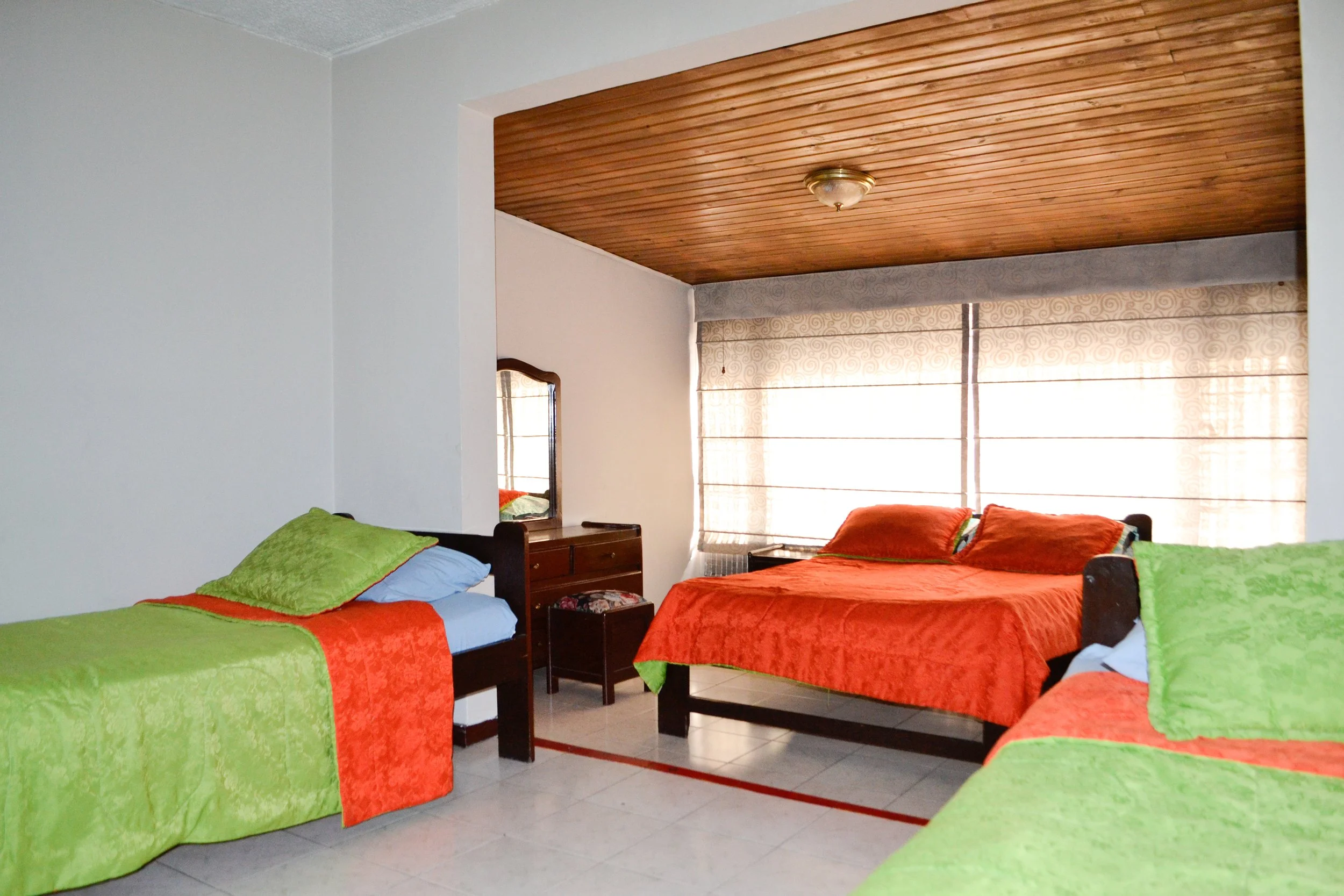 Habitación con tres camas con cobertores verdes y naranjas, dos ventanas con cortinas claras, un tocador de madera con espejo, y suelo de baldosa blanca.