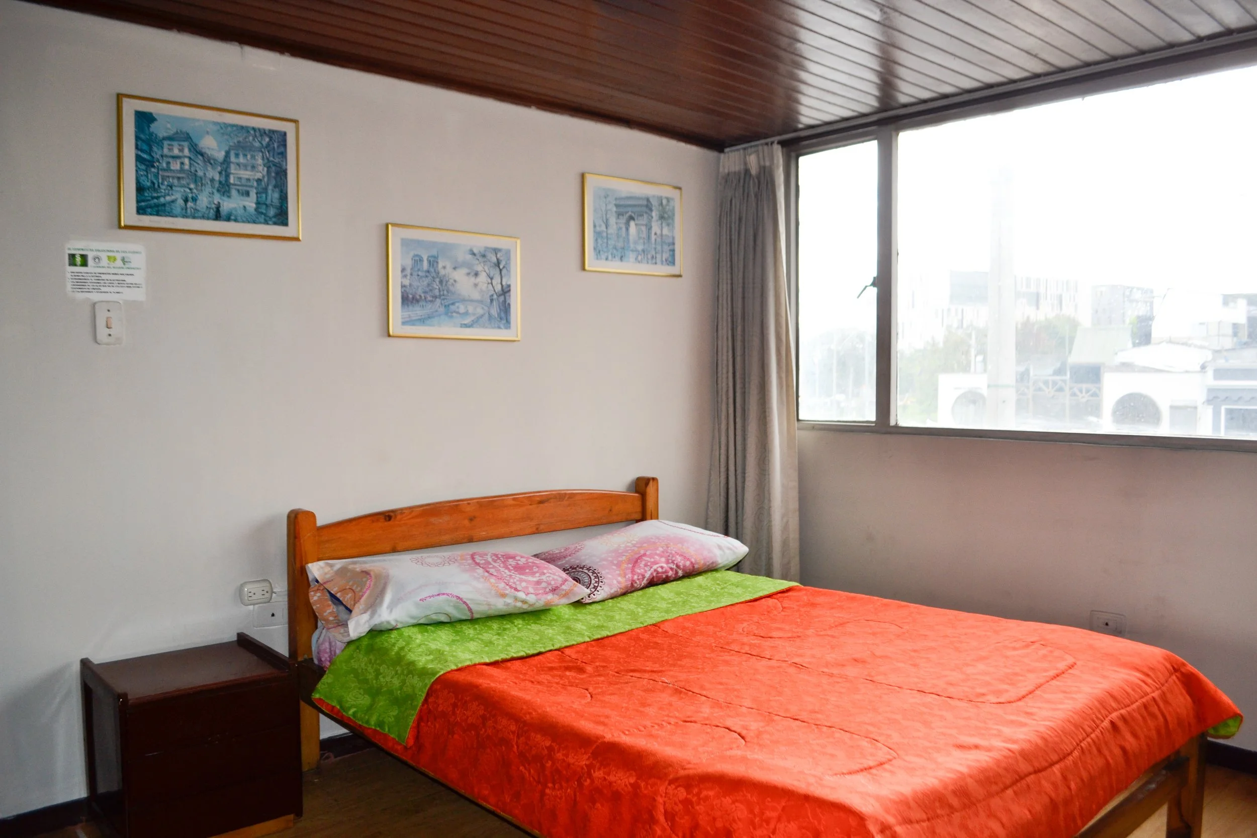 Habitación con cama de madera, cobertor naranja, almohadas, ventana grande con cortinas, cuadros en la pared.