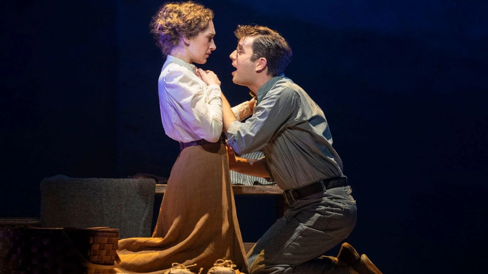 A man kneeling and a woman standing close, looking intensely at each other, in a theatrical scene with a dark background.