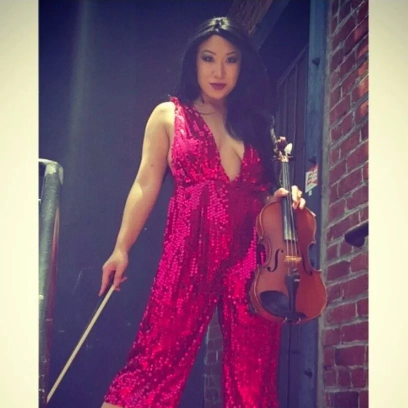 Woman in a sparkly red dress holding a violin and a bow, standing against a brick wall.