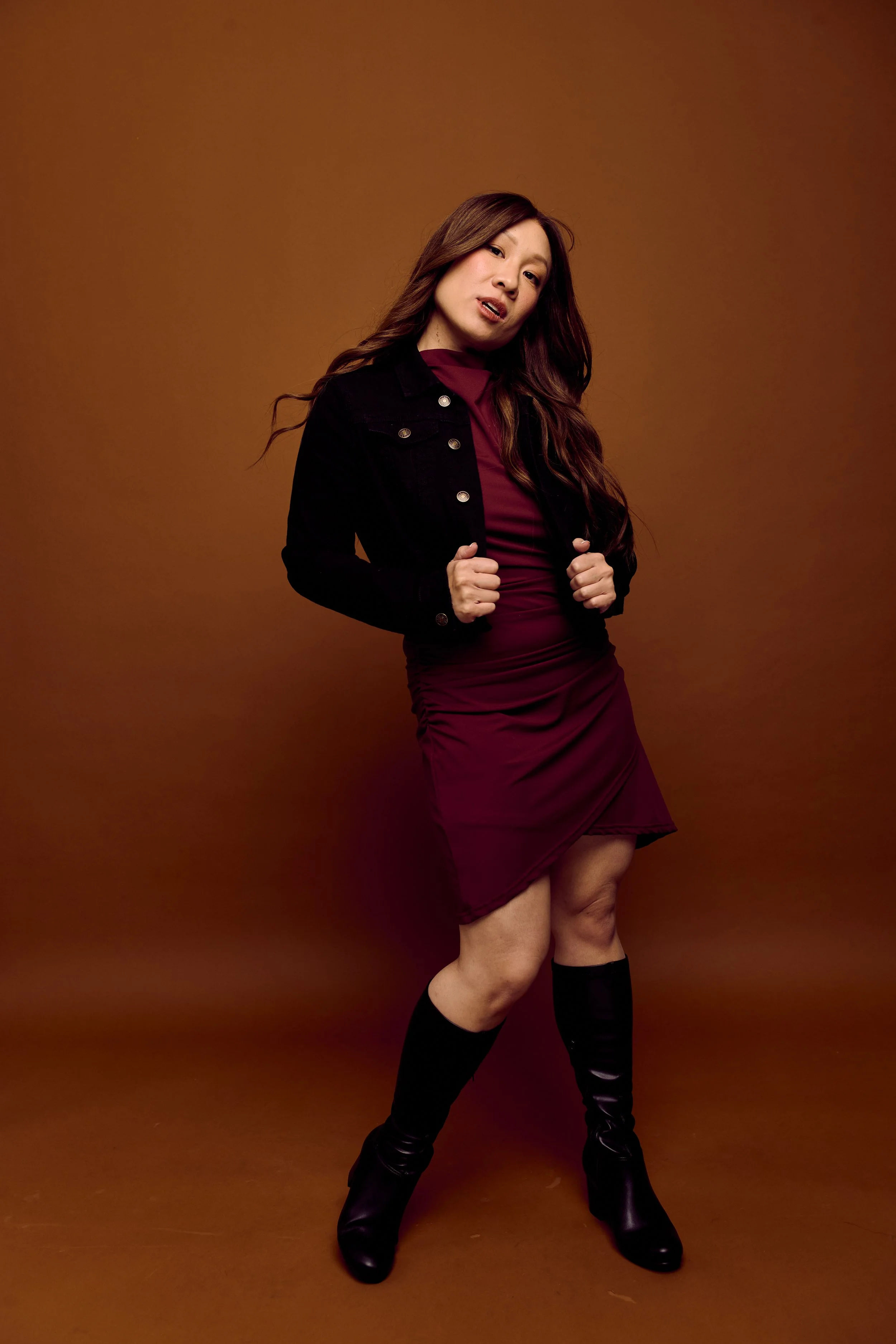 Tina Golden Picture. Musical Theater Performer. A woman with long brown hair wearing a black jacket, a burgundy dress, and knee-high black boots standing against a brown background.