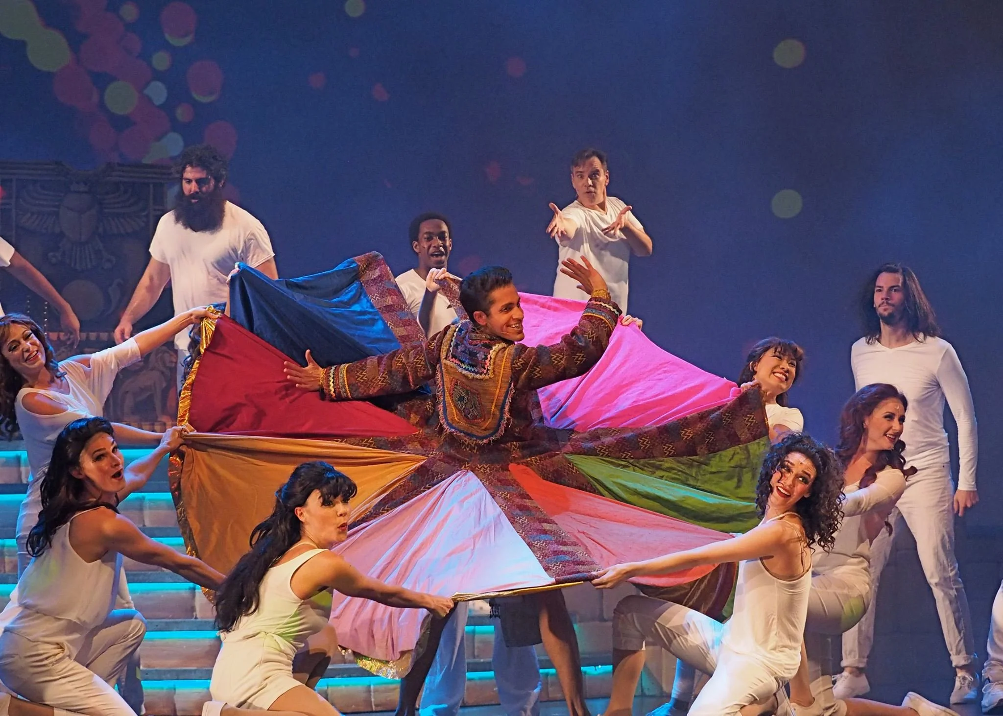 Group of performers on stage participating in a theatrical dance, with one man in the center holding a colorful fabric, surrounded by others, some kneeling and some standing, with a dark background and stage lighting.