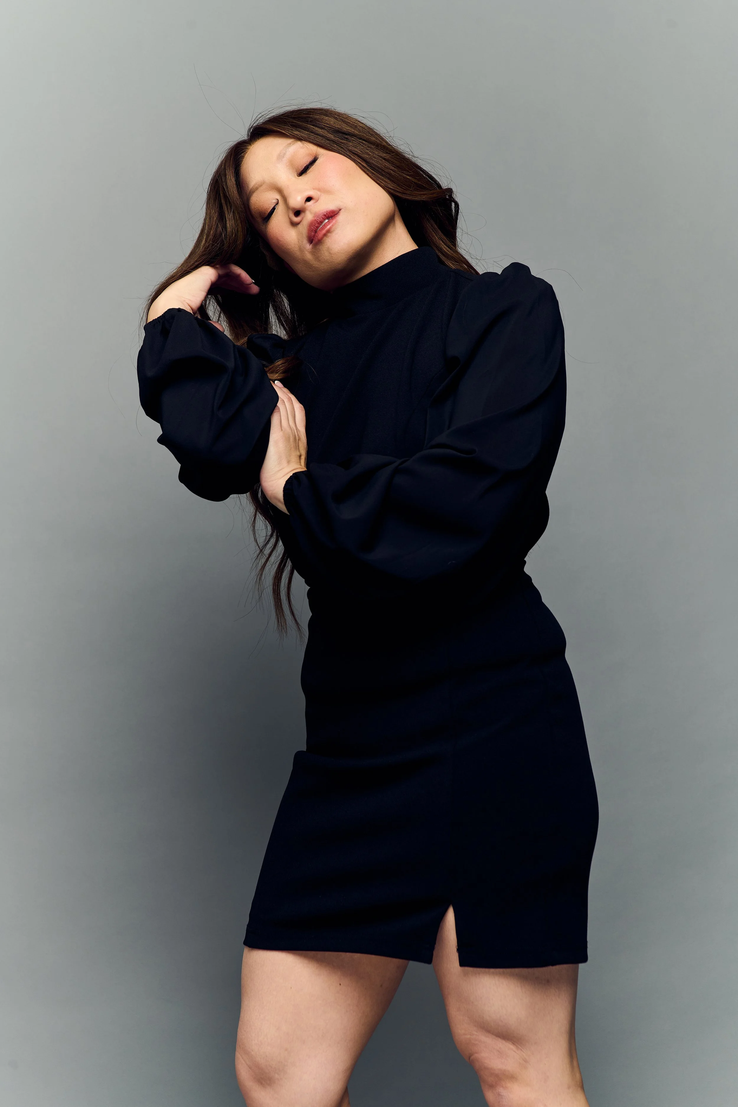 Tina Golden Picture. Musical Theater Performer. A woman with long brown hair, wearing a black dress, is posing with her eyes closed and smiling softly against a plain gray background.