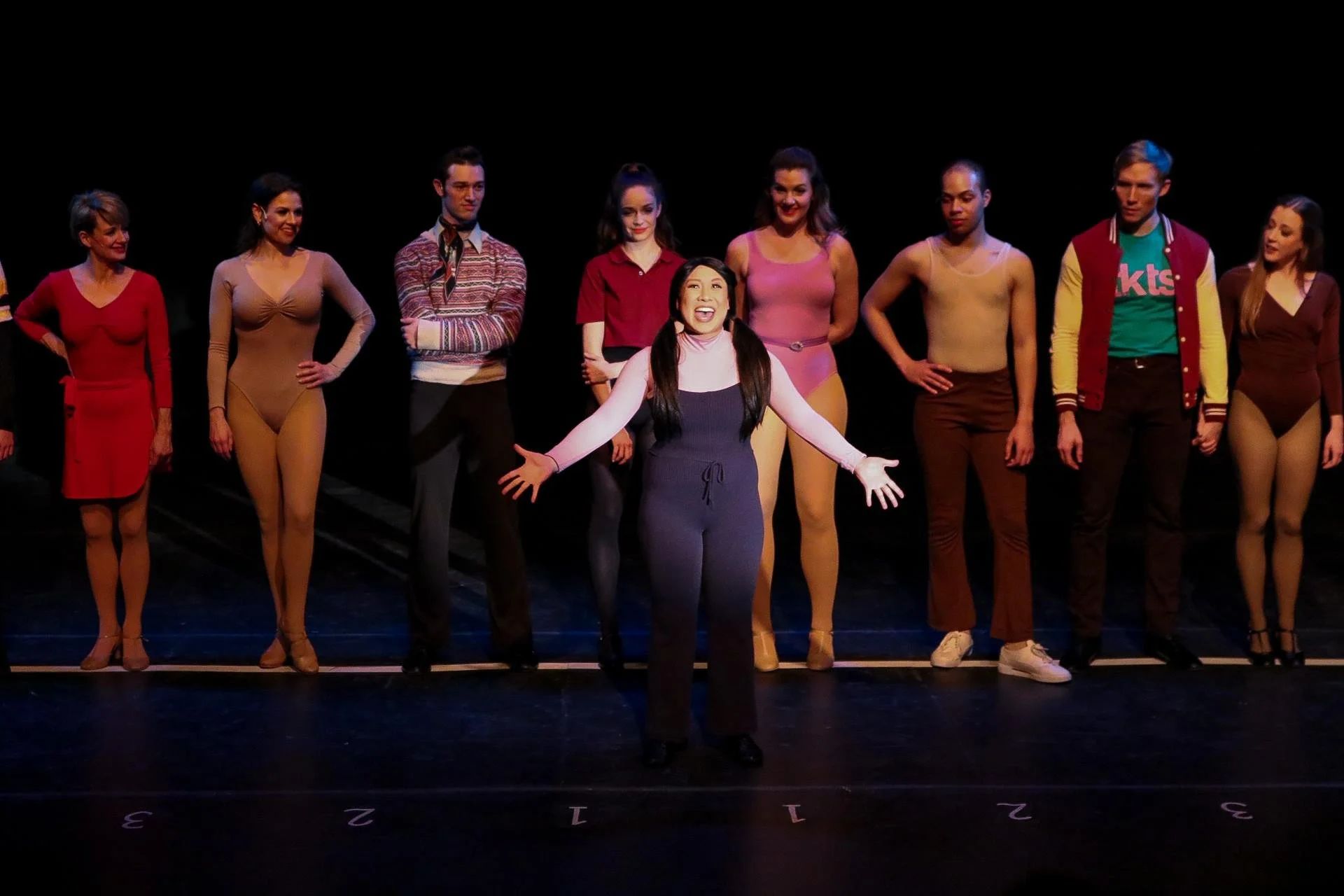 Tina Golden in A Chorus Line with pigtails standing center stage, gesturing with open arms and smiling, surrounded by a diverse group of performers in colorful costumes, on a black stage with numbered lines.
