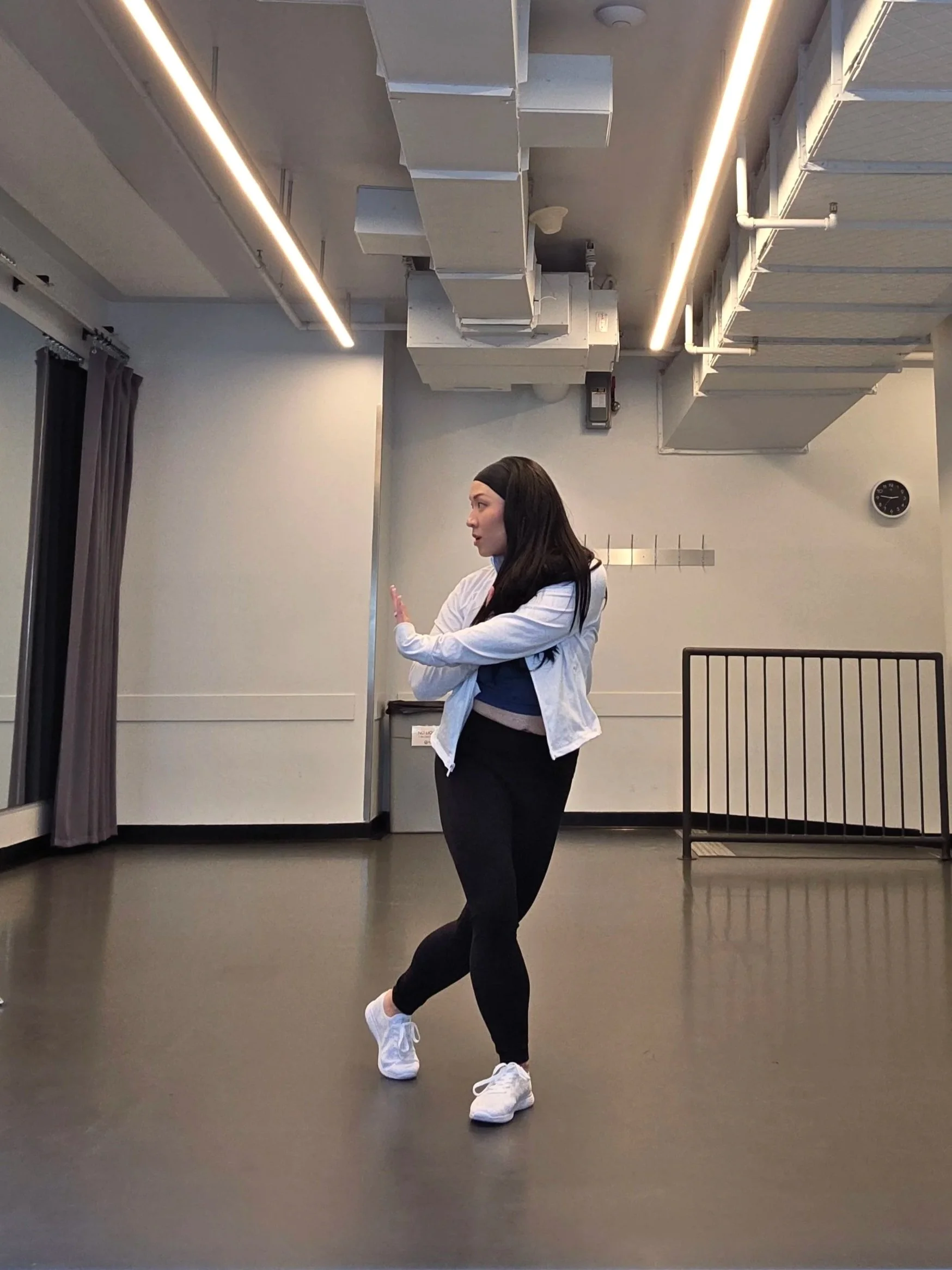 Tina Golden. A woman in black leggings and white sneakers in a dance studio, standing in a pose with one arm extended and the other bent, with her head turned to the side.