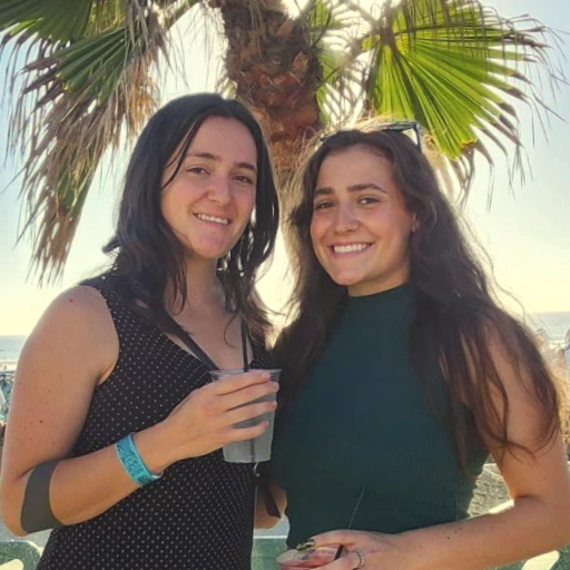 Women smiling and holding cocktails at an event with a palm tree in the background during sunset.