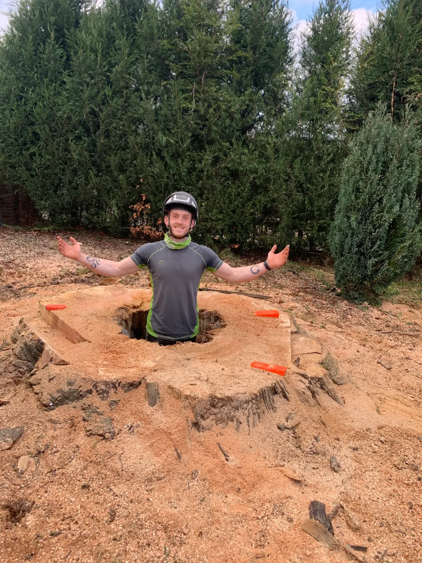 A man in a helmet and athletic clothing standing in a large tree stump, with his arms outstretched and smiling, surrounded by greenery in an outdoor setting.
