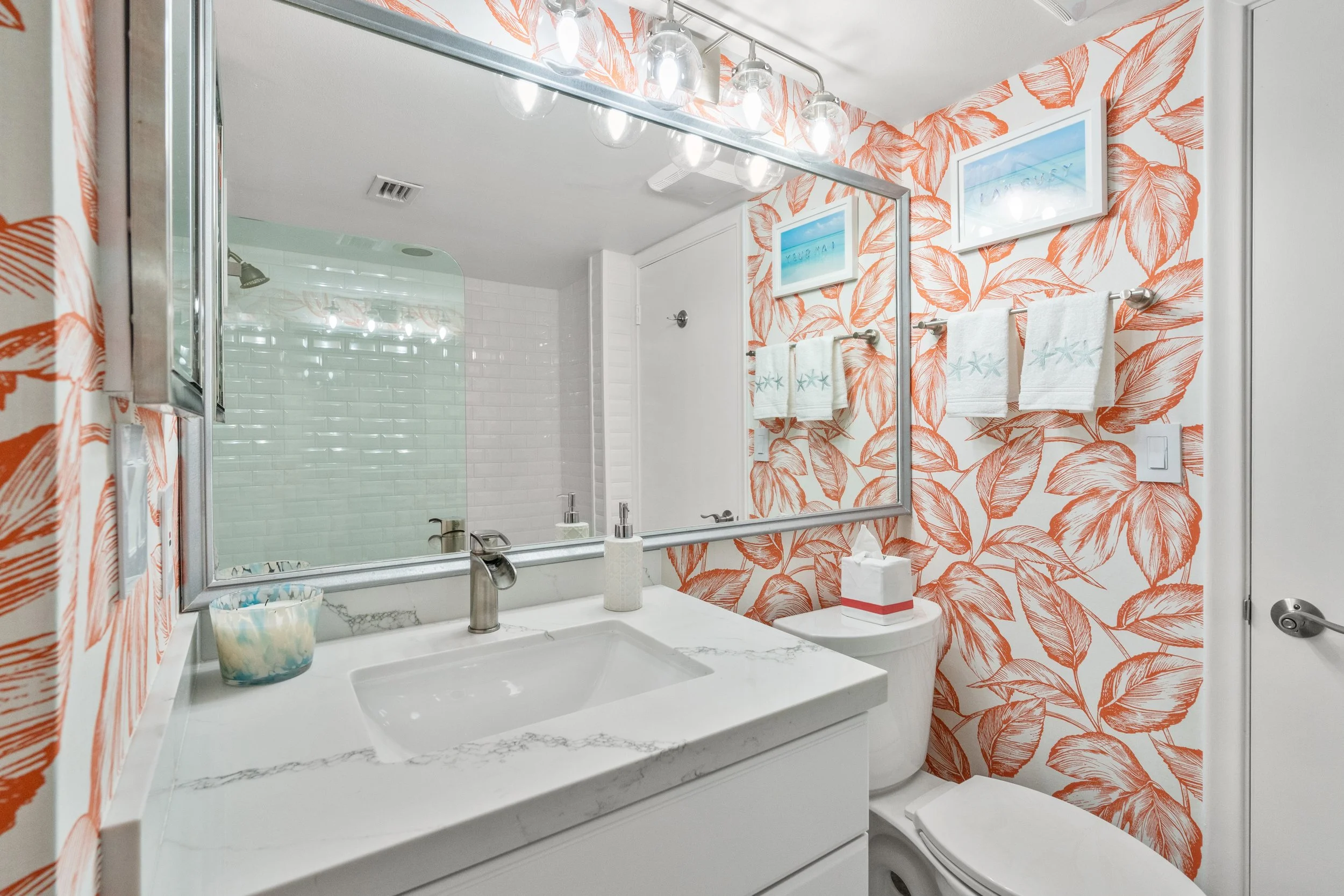 Bathroom with orange and white leaf-patterned wallpaper, large mirror, white marble countertop sink, white toilet, and framed artwork near ceiling