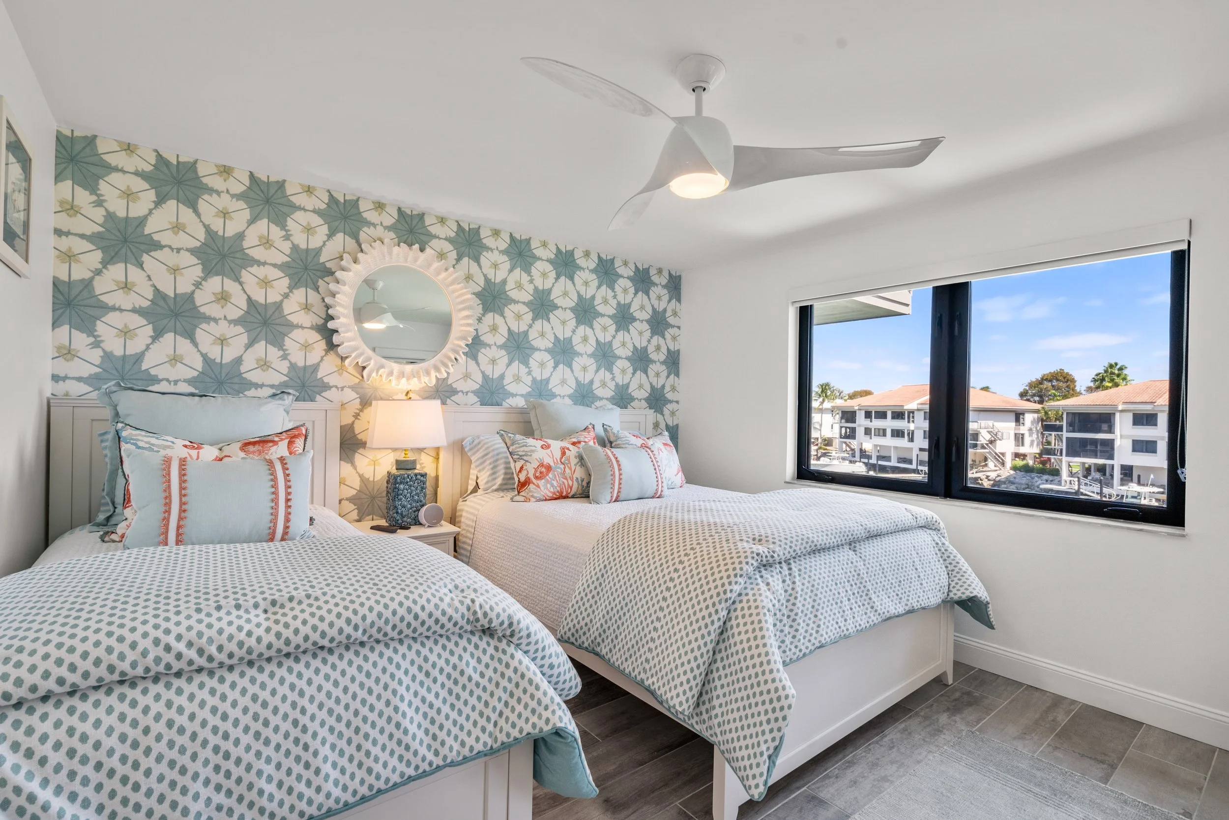 A bright bedroom with two twin beds, patterned blue and white bedding, decorative pillows, a nightstand with a lamp, a round mirror on a floral wallpapered wall, a ceiling fan, and a window showing a view of neighboring buildings and a blue sky.