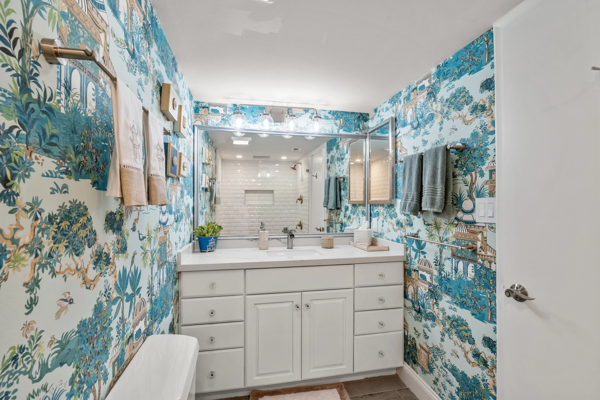 Bathroom with wallpaper featuring a blue and beige scenic pattern, white vanity with a marble top, large mirror, gray towels on a stainless steel rack, and a shower area with white subway tiles and brass fixtures.