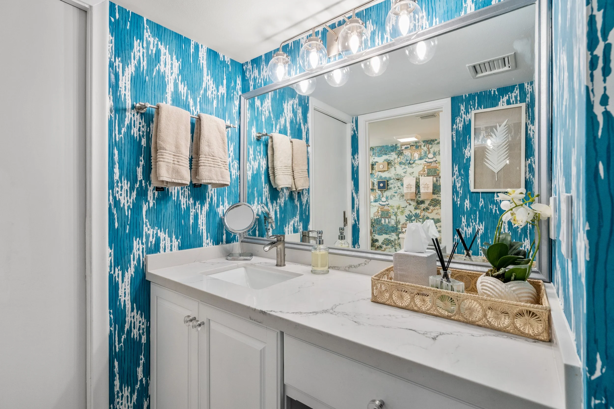 Bathroom vanity with a white marble countertop, silver faucet, mirror, beige towels, and decorative tray with tissues, scented reeds, and a small plant. Blue and white patterned wallpaper on the walls.