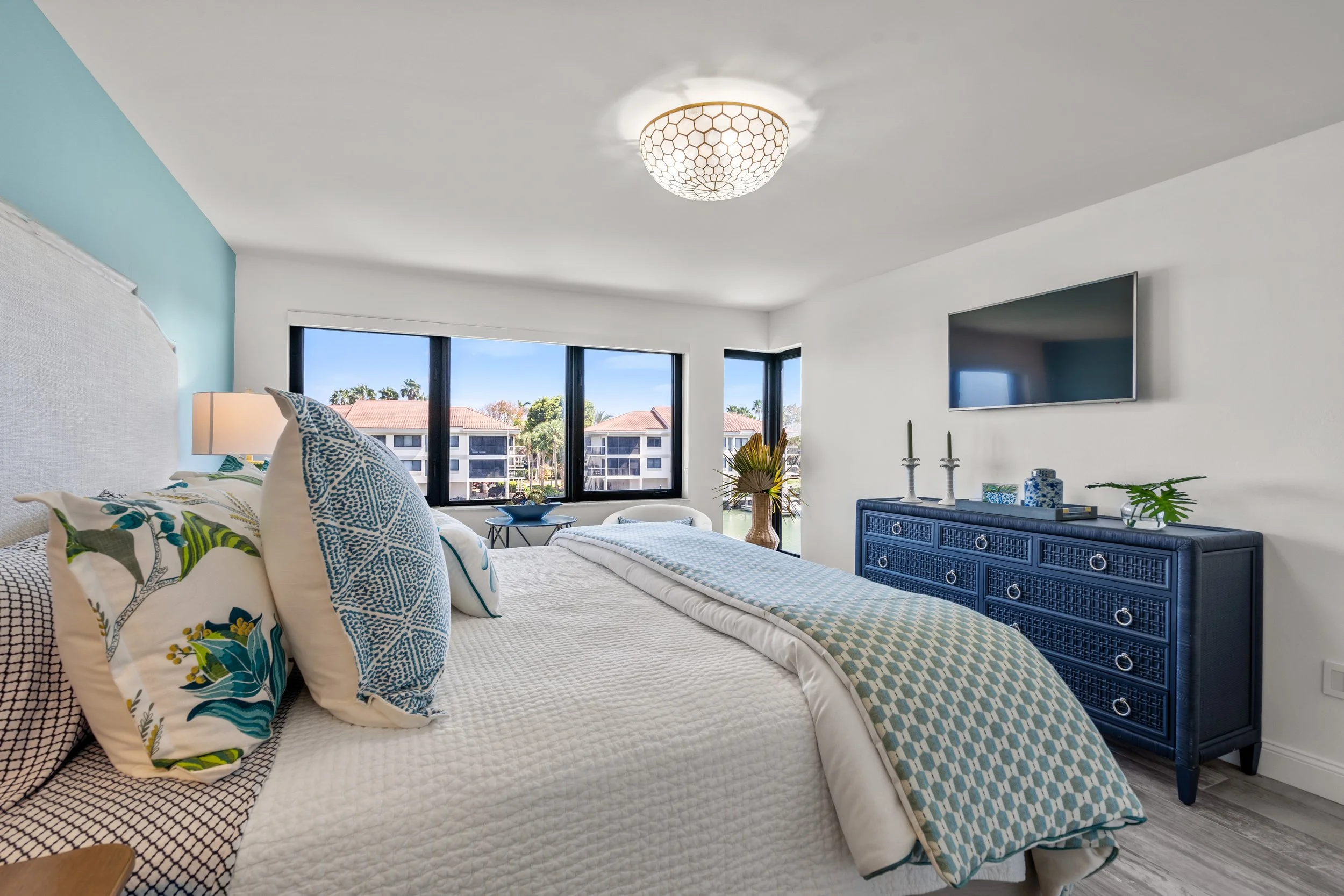 Bright, modern bedroom with white walls, a large bed with patterned pillows and a quilt, a blue dresser, a wall-mounted TV, and large windows showing a view of neighboring buildings and trees.