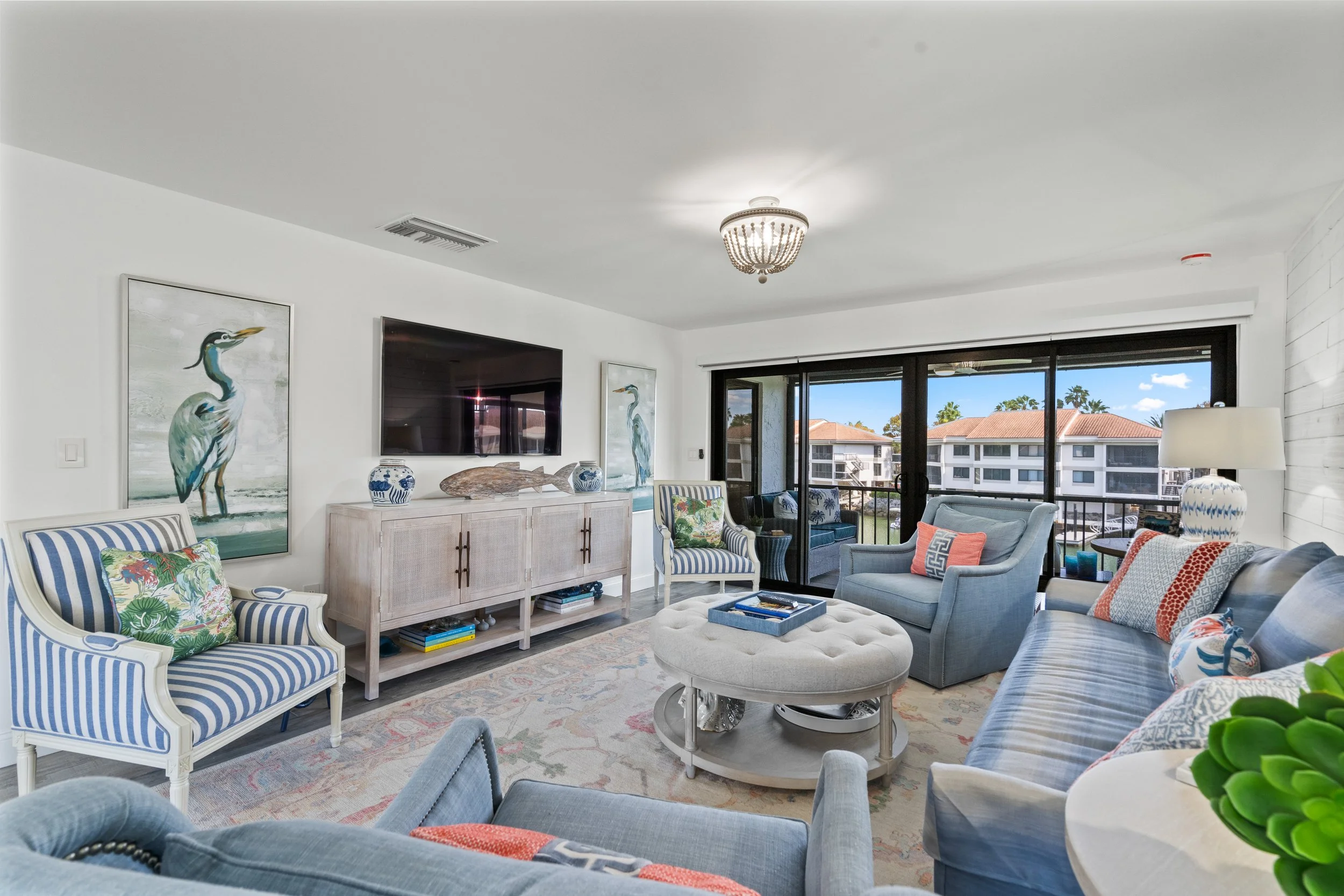 Living room with blue and white furniture, tropical decor, and large sliding glass doors leading to a balcony with outdoor seating and a view of neighboring buildings.