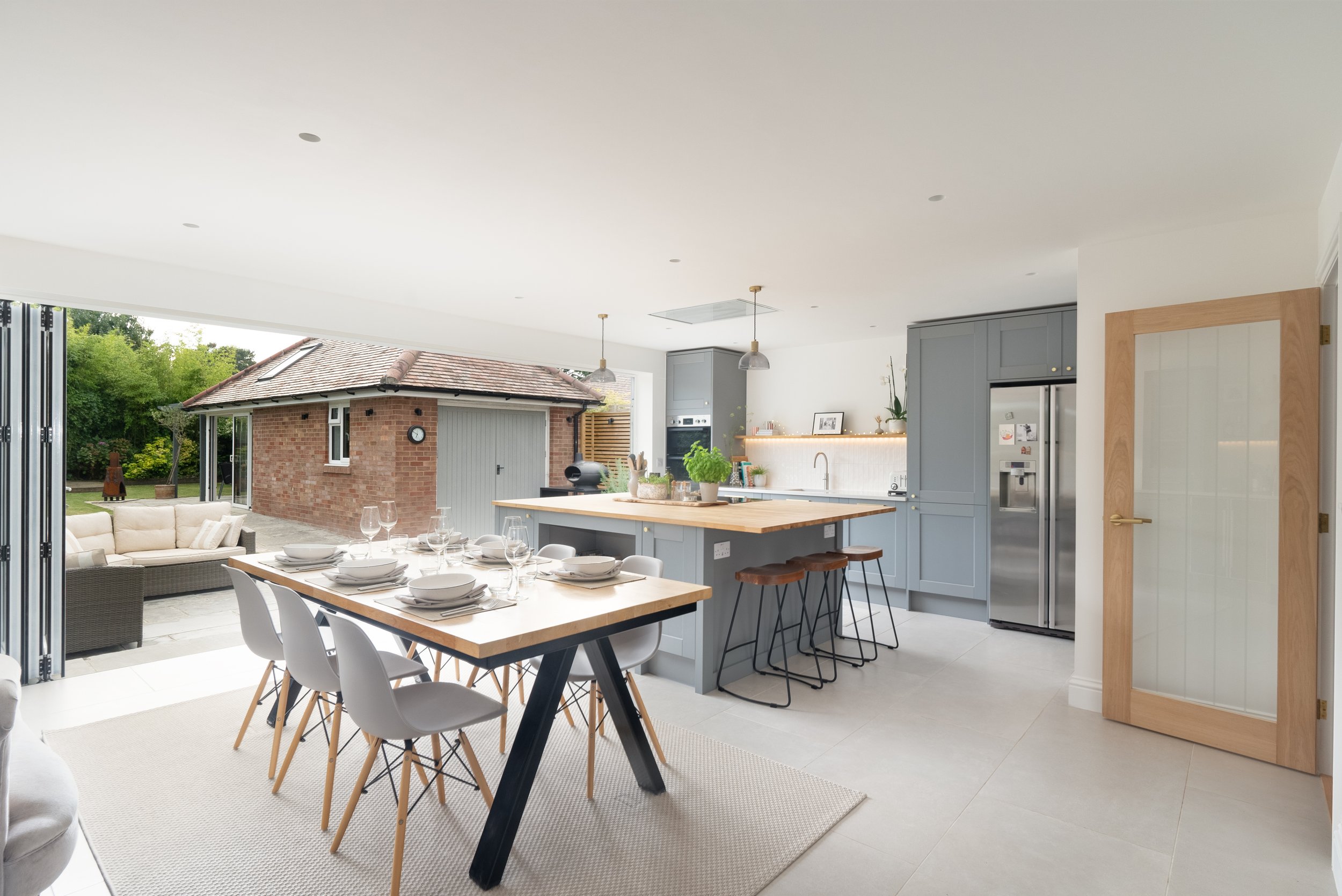 Open-plan kitchen and dining area with a wooden dining table set for six, a kitchen island with bar stools, gray cabinetry, and a view of an outdoor patio with seating and a small brick building.