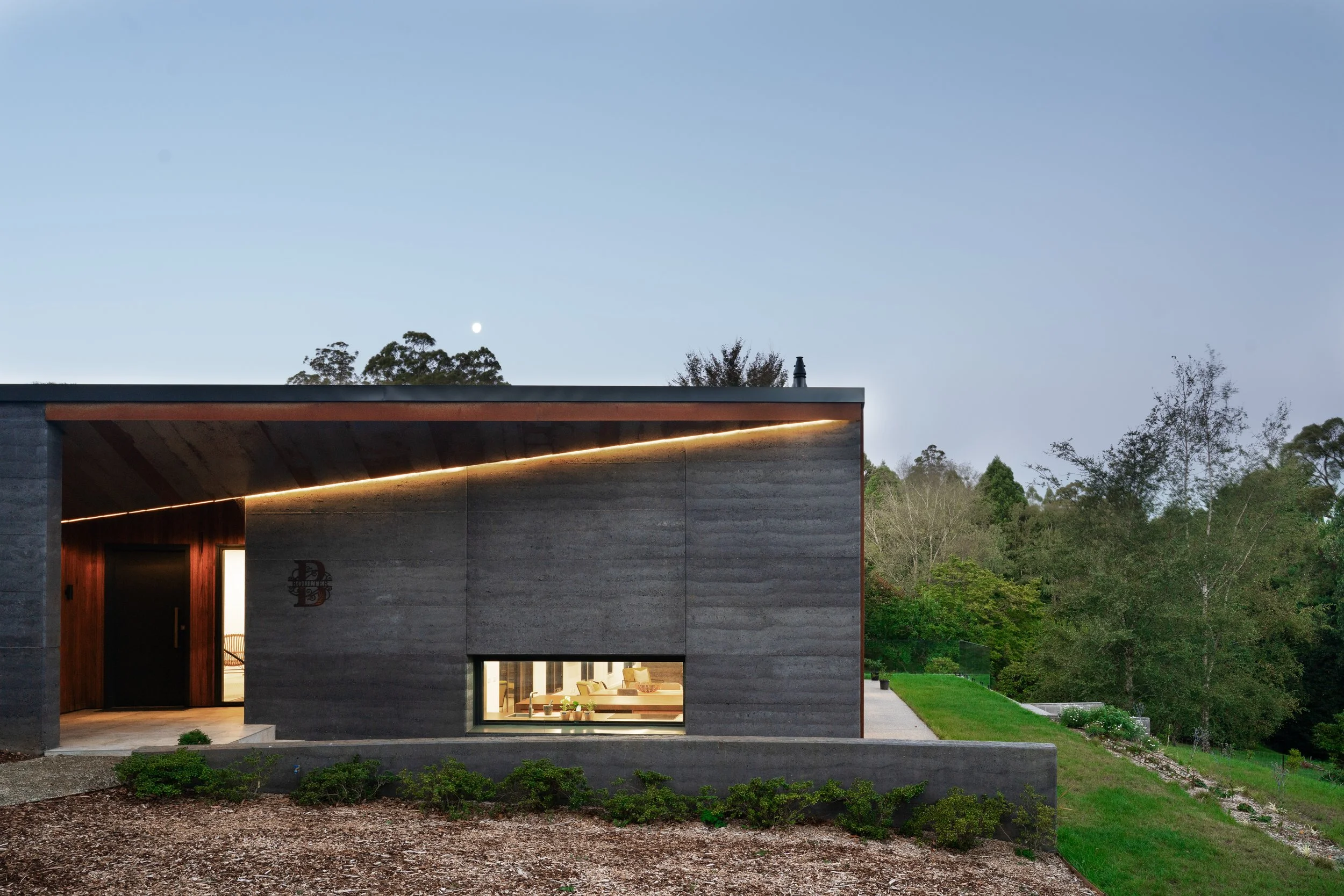 Modern house with concrete exterior, a large window showing a living room, surrounded by a lawn and trees, under a clear sky with the moon visible.