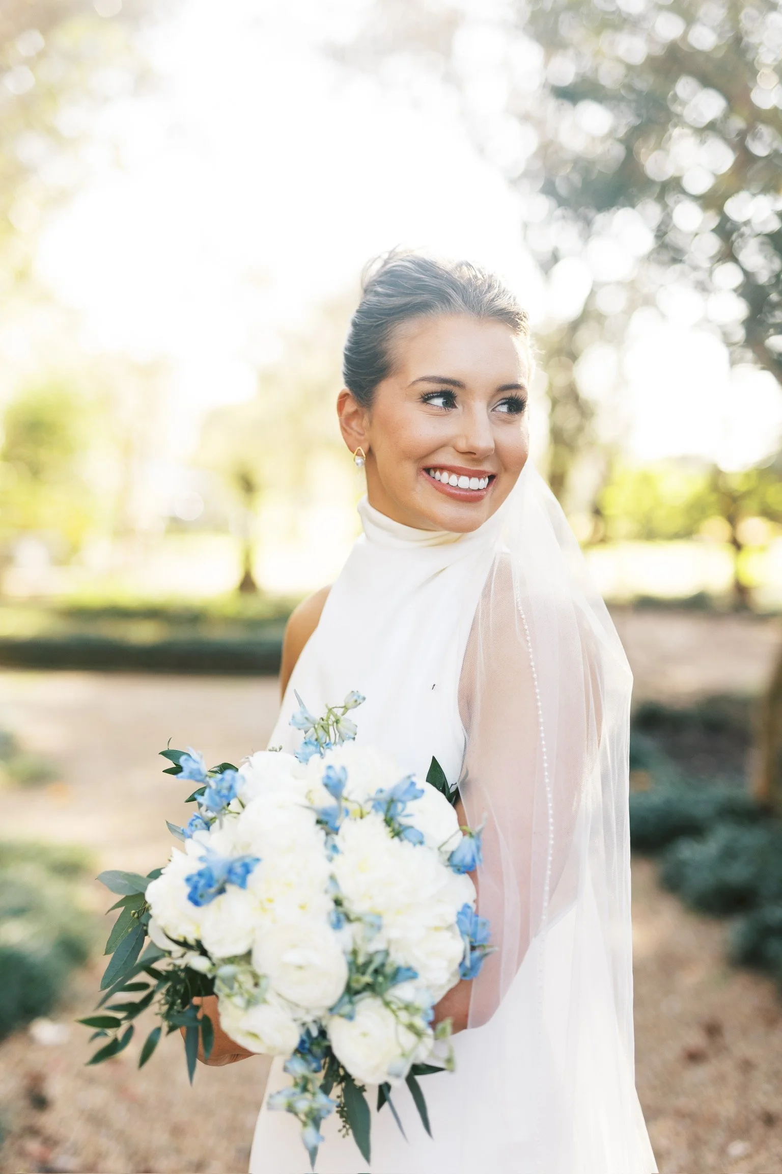 A smiling bride holding a bouquet of white and blue flowers outdoors on a sunny day.