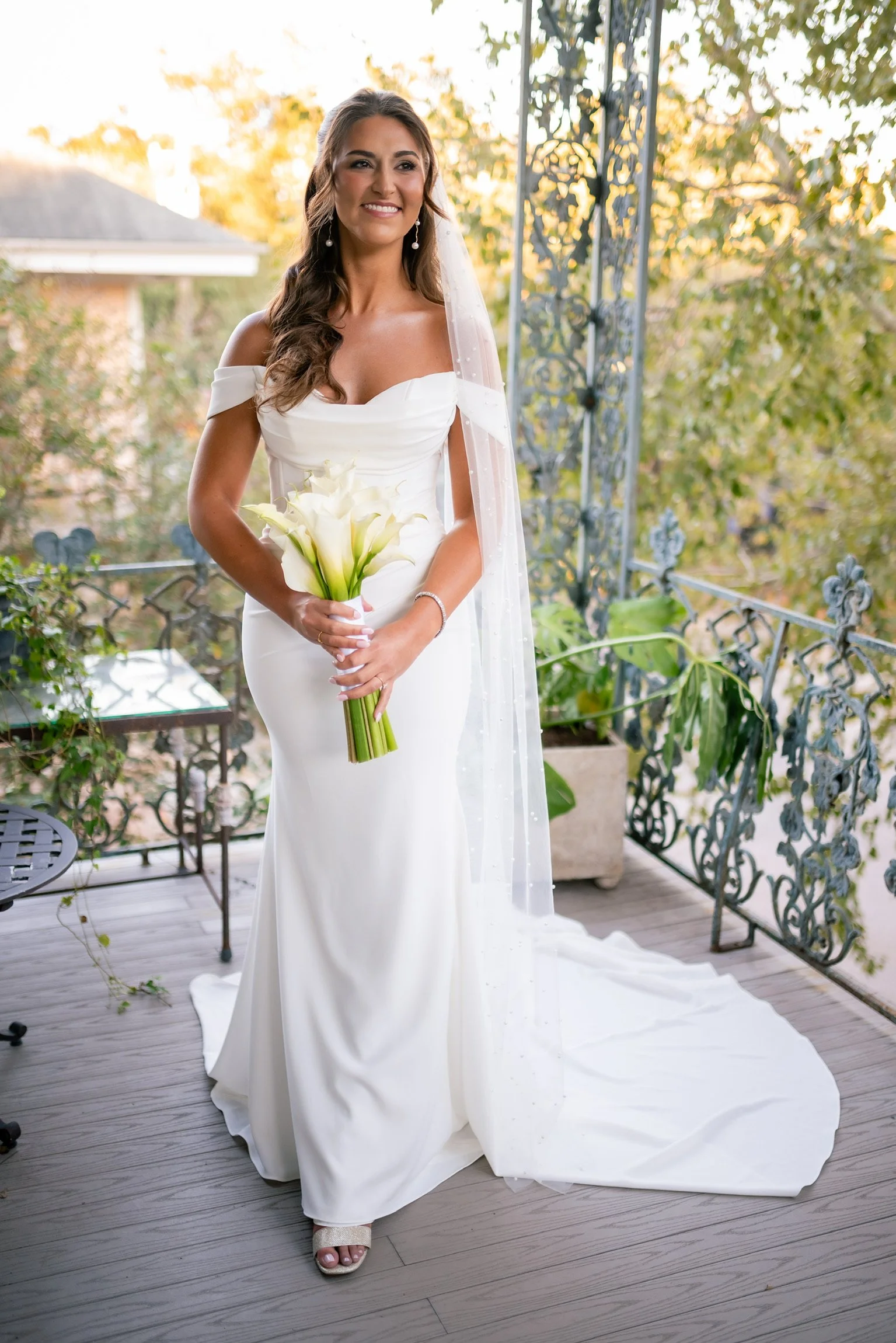 A woman in a white wedding dress standing on a balcony holding a bouquet of white calla lilies, with trees and sunset in the background.