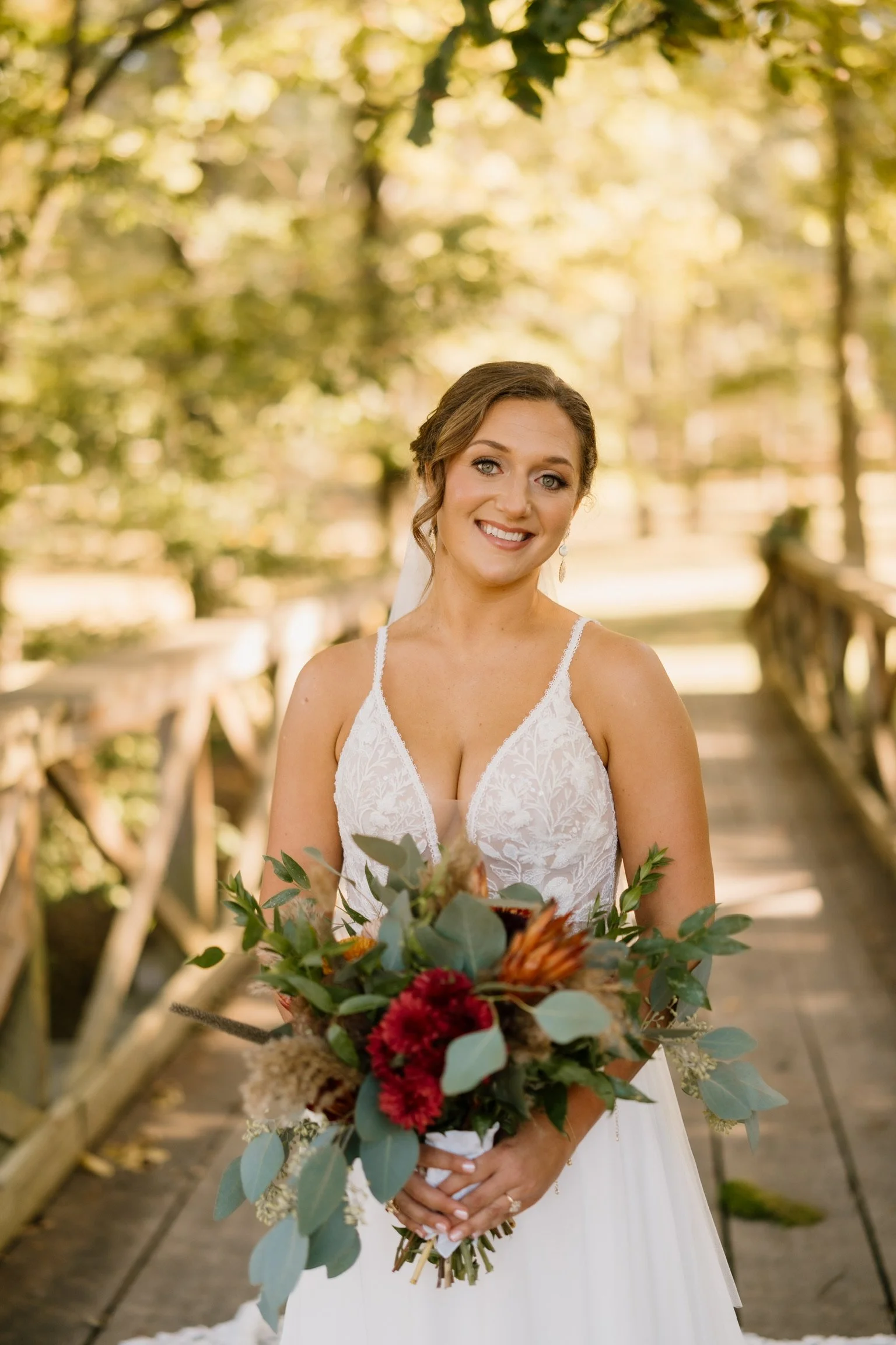A bride in a white lace wedding dress holding a bouquet of flowers on a wooden bridge surrounded by trees.