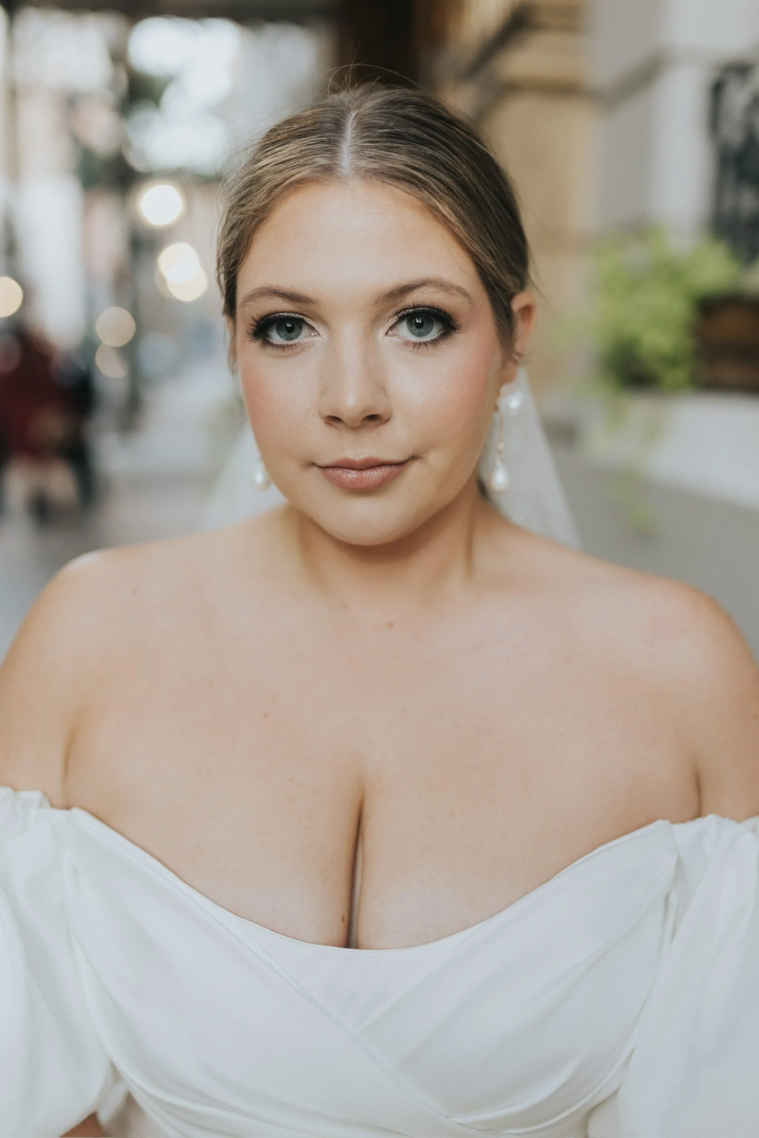 A portrait of a bride with blue eyes and light brown hair in an updo, wearing pearl earrings and a white off-shoulder wedding gown.