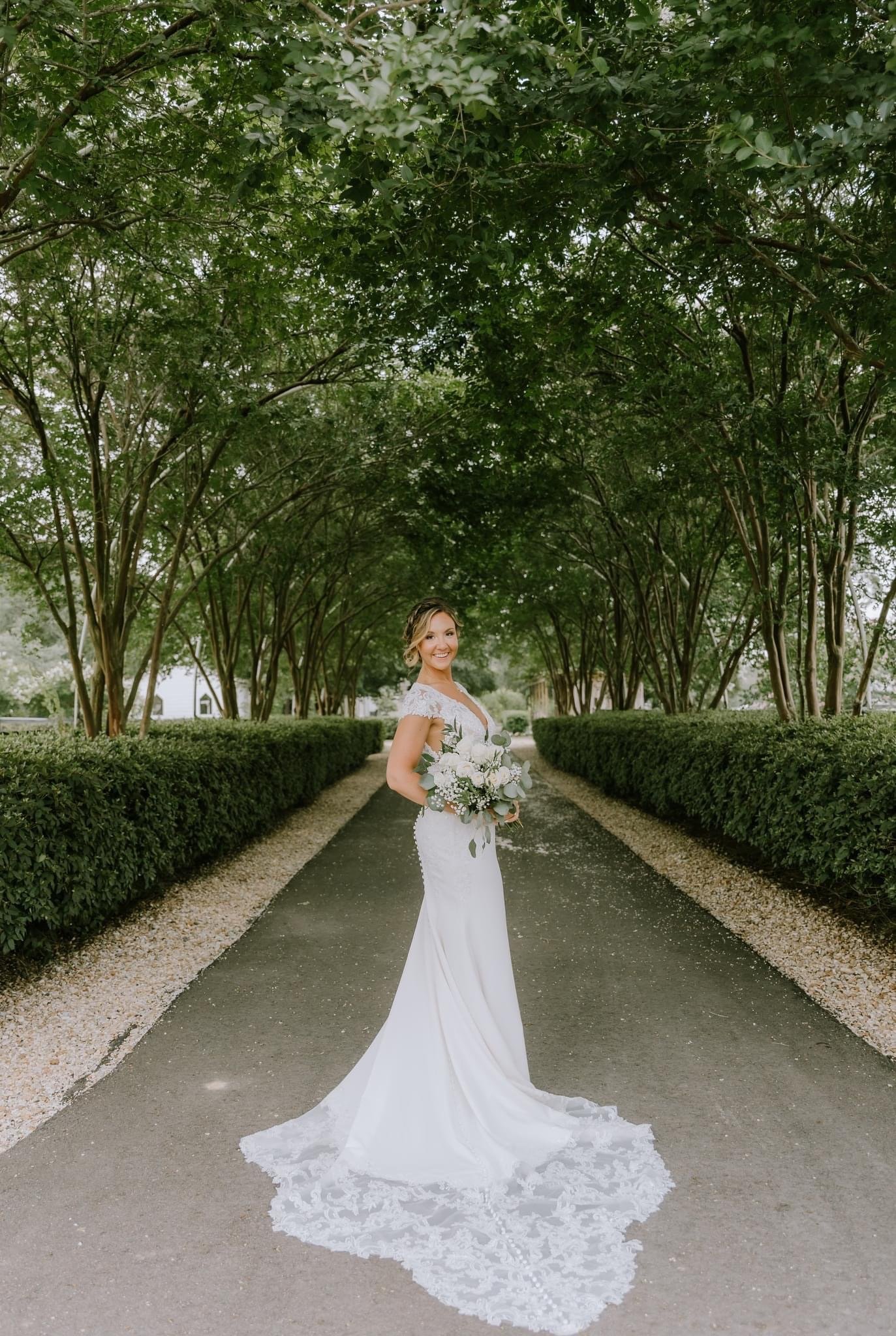 A bride in a white wedding dress holding a bouquet of white flowers standing on a tree-lined pathway.
