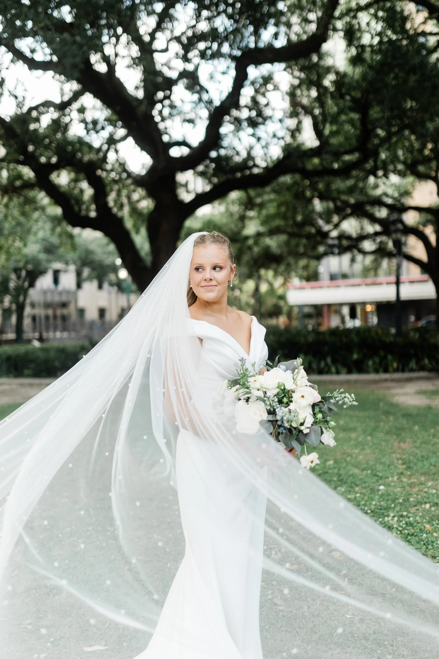 Bride in a white wedding dress holding a bouquet of white flowers and greenery, standing outdoors in front of a large tree with sprawling branches.