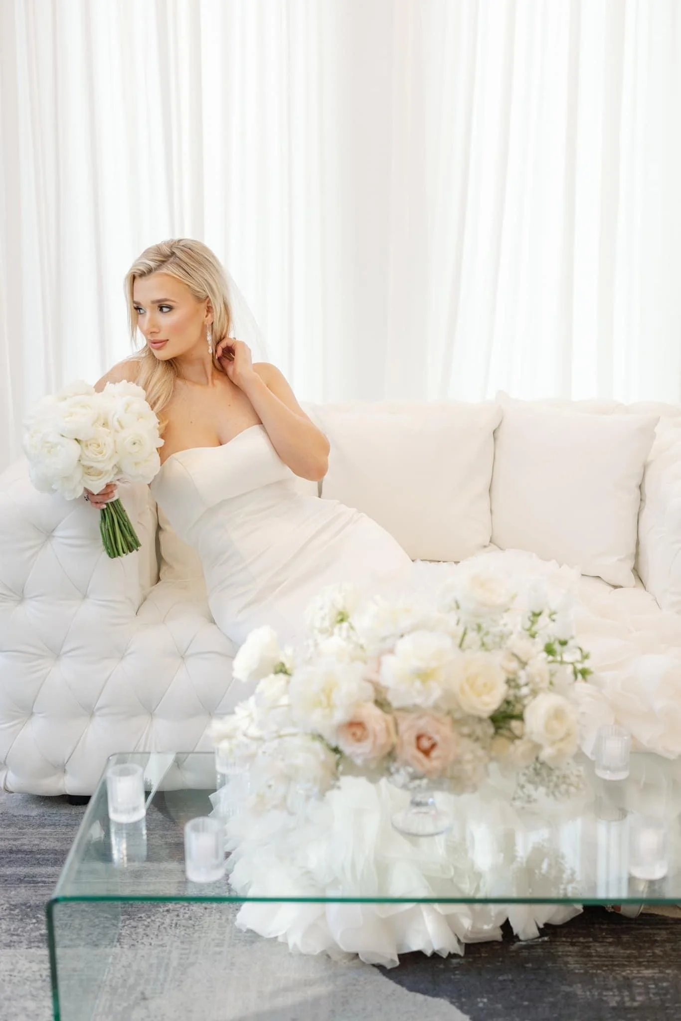 A bride in a strapless white wedding gown sitting on a white tufted sofa, holding a bouquet of white roses, with floral arrangements on a glass coffee table in front of her, and sheer white curtains in the background.
