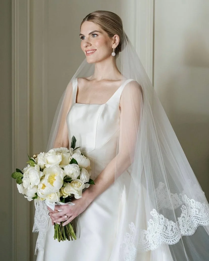Bride in white wedding dress holding a bouquet of white flowers, wearing a veil and earrings, standing indoors with a neutral background.