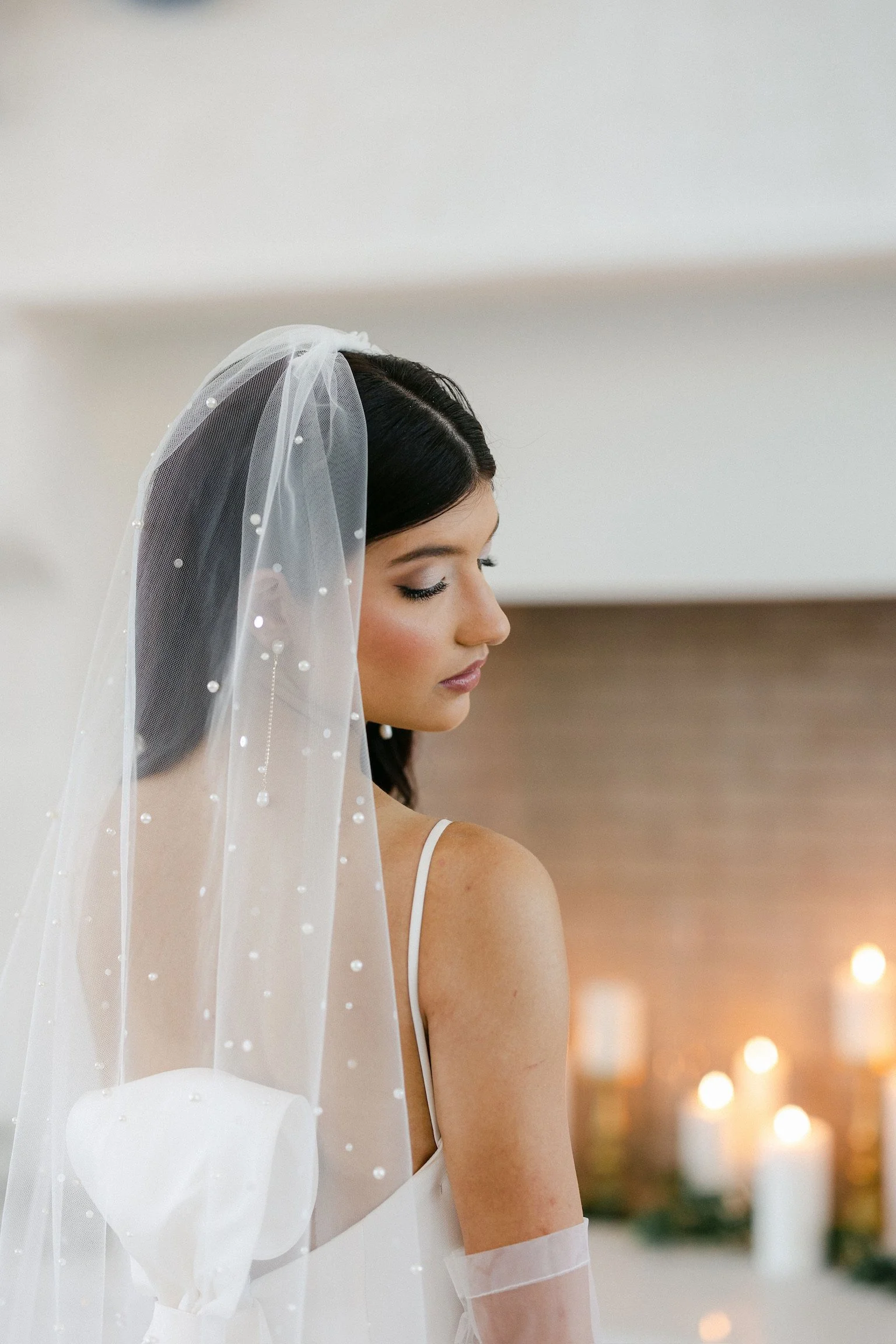 A bride with dark hair in a white dress and veil standing with her eyes closed, soft lighting, candles in the background.