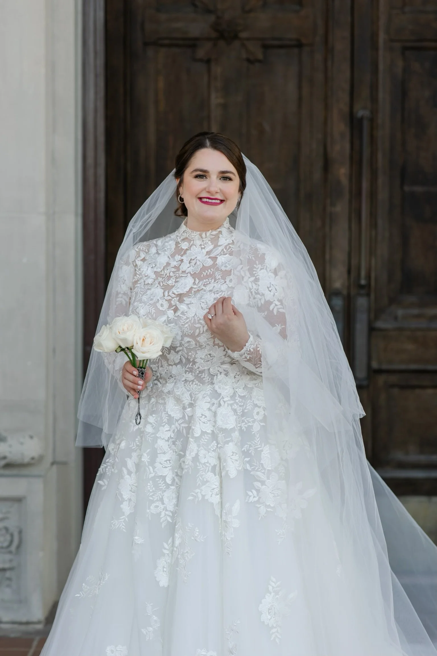 Bride in a lace wedding dress holding a bouquet of white roses, standing in front of a wooden door.