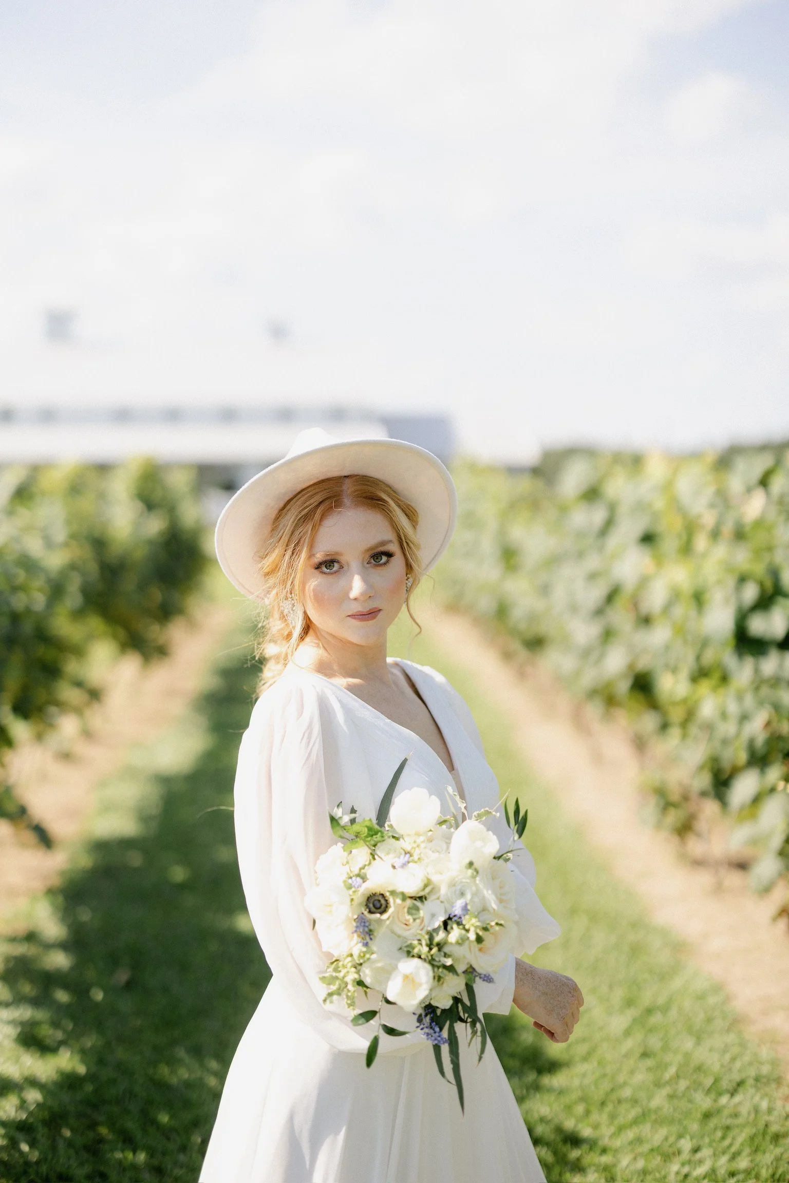 A woman in a white dress and wide-brimmed hat holding a bouquet of white flowers standing in a vineyard.