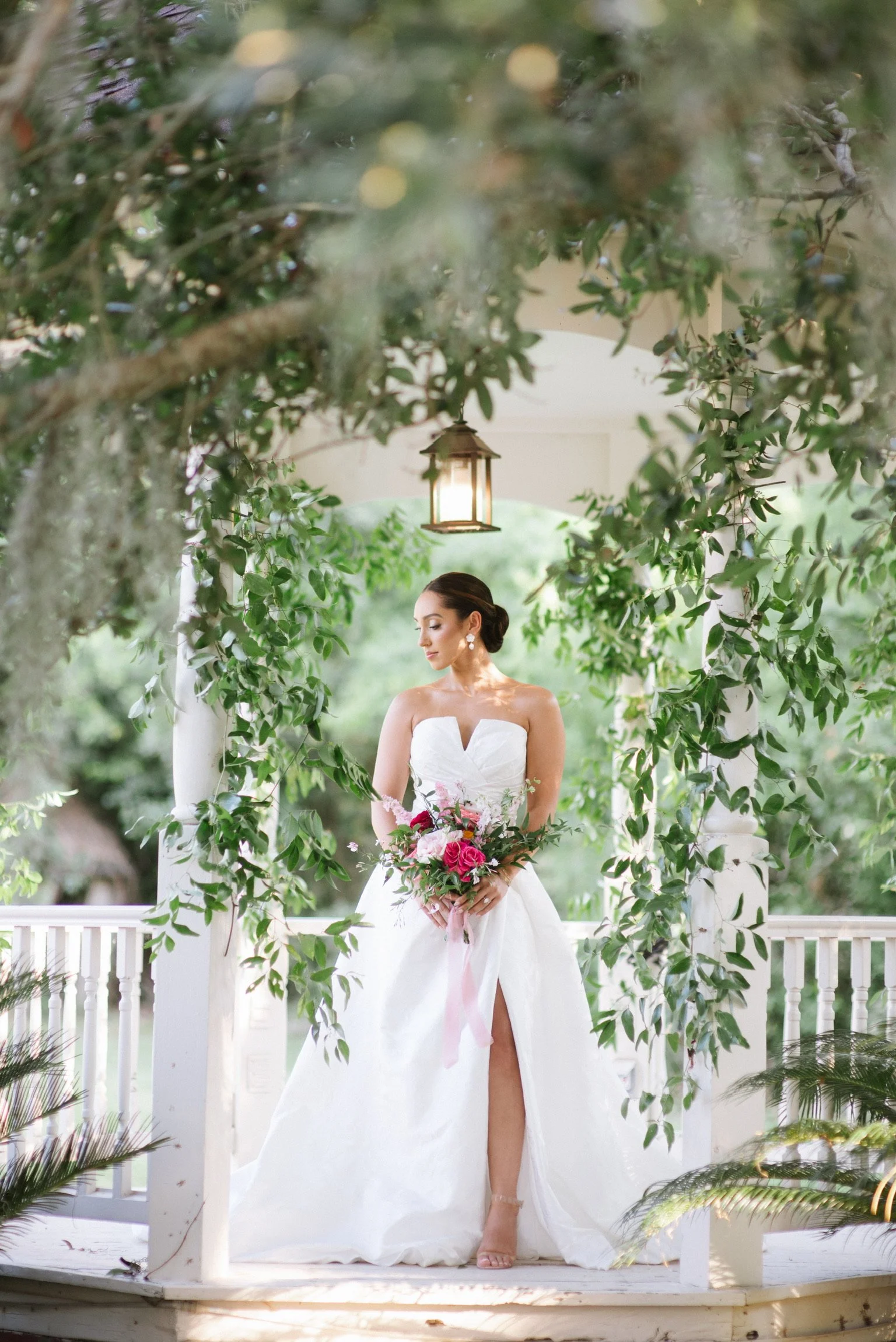Bride in a white wedding dress holding a bouquet, standing on a gazebo decorated with greenery and a hanging lantern, outdoors.