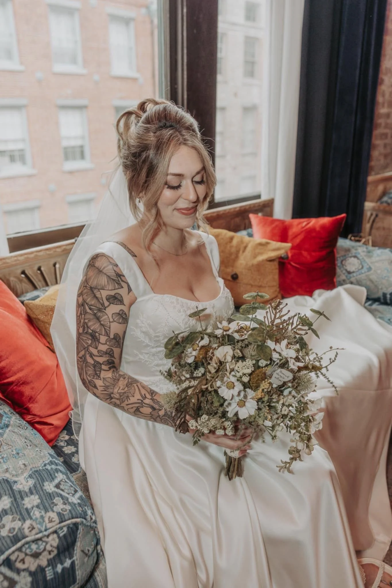 A bride with tattoos on her arms, wearing a white wedding dress and veil, sitting on a sofa with orange and patterned cushions near a window, holding a bouquet of flowers.