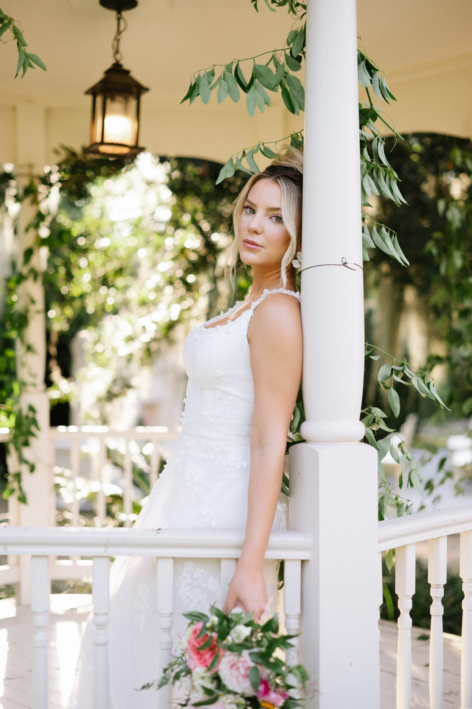 A bride in a white wedding dress holding a bouquet, leaning against a white railing on a porch decorated with greenery, with a lantern hanging above.