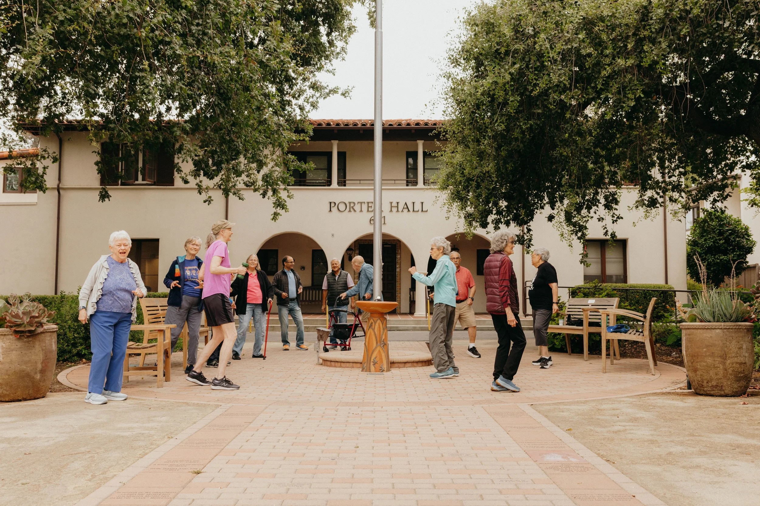 A group of older adults gather and socialize outdoors in a courtyard at Pilgrim Place, walking, talking, and moving together in front of Porter Hall, with benches, trees, and the campus buildings in the background.