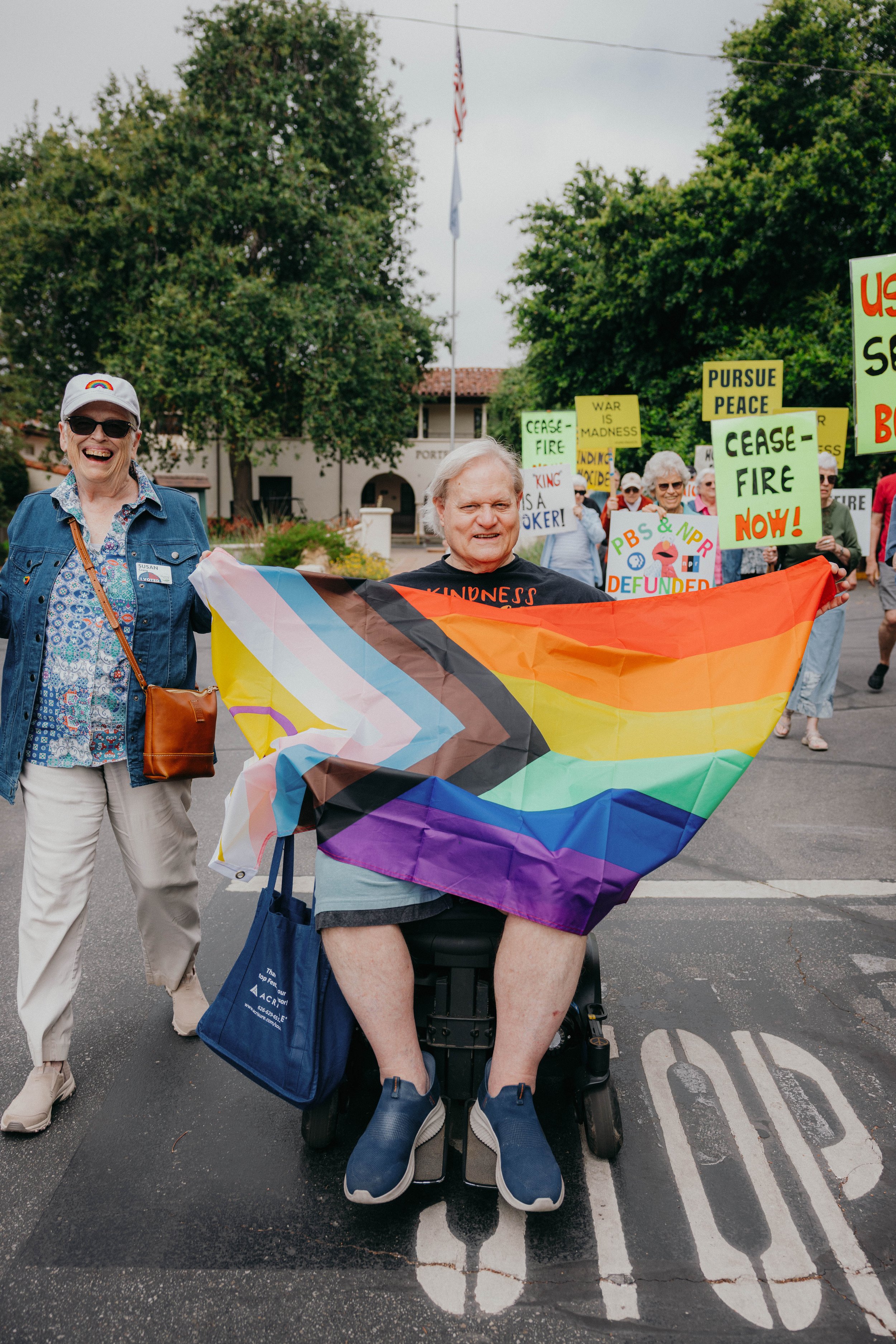 Two older adults from Pilgrim Place participate in a Pride march, smiling and holding a rainbow progress flag; one person uses a mobility scooter while the other walks beside them, with protest signs and a community setting in the background.