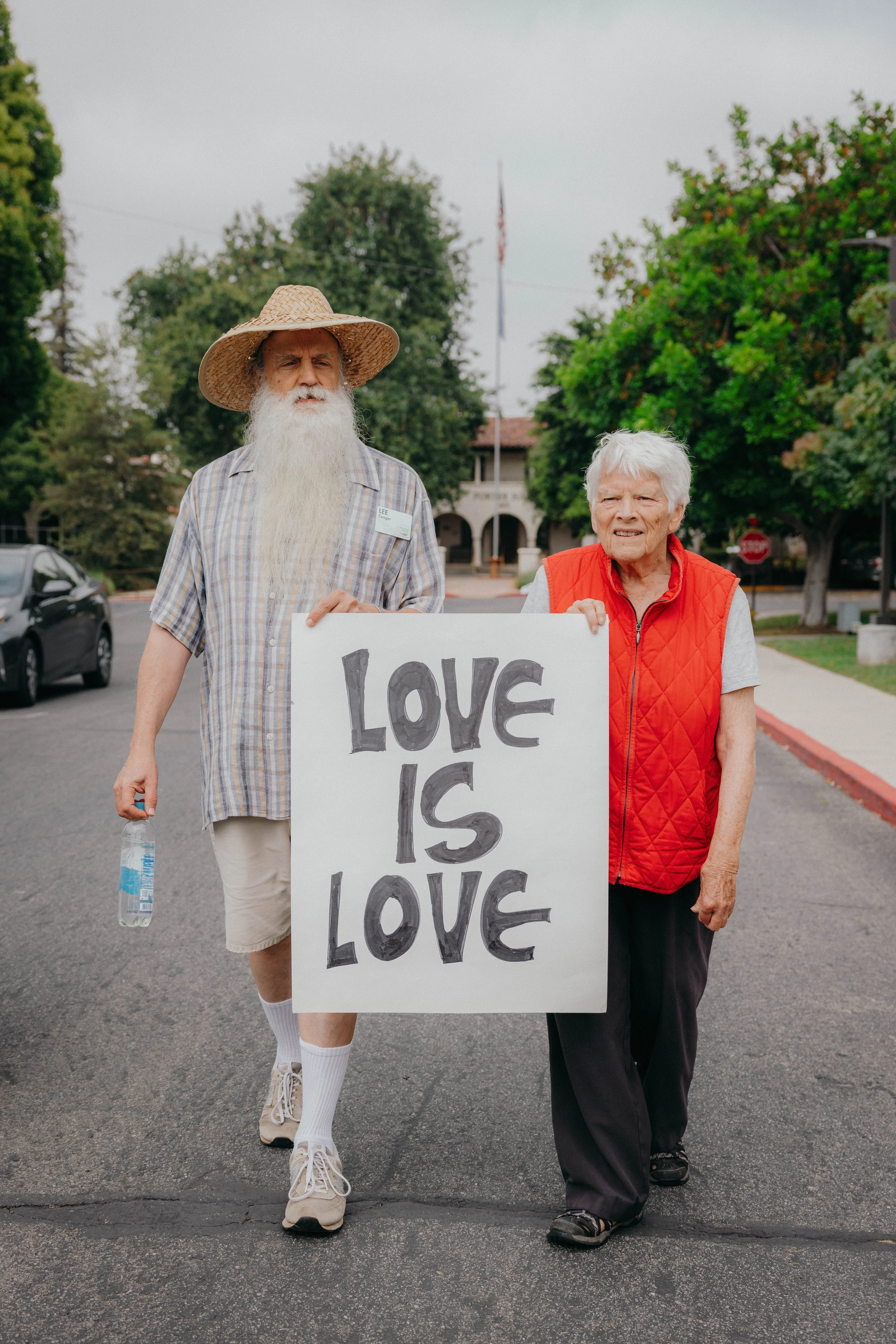 Two older adults stand together on a campus street holding a hand-lettered sign that reads “Love Is Love,” conveying support for LGBTQ+ inclusion and affirming values within a retirement community setting.