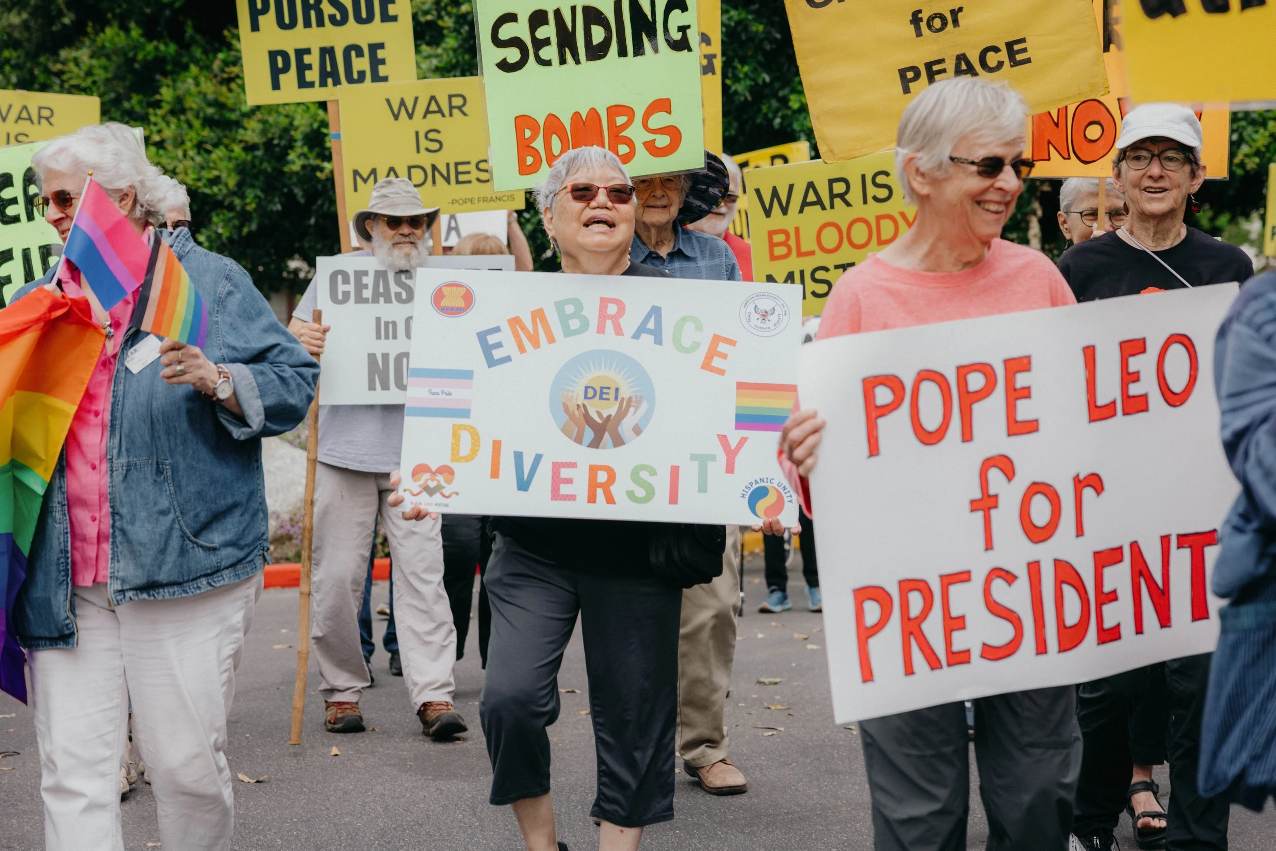 A group of older adults march together at a peace and justice rally, smiling and holding signs that read “Embrace Diversity,” “Pursue Peace,” and other messages supporting inclusion, LGBTQ+ pride, and social justice.