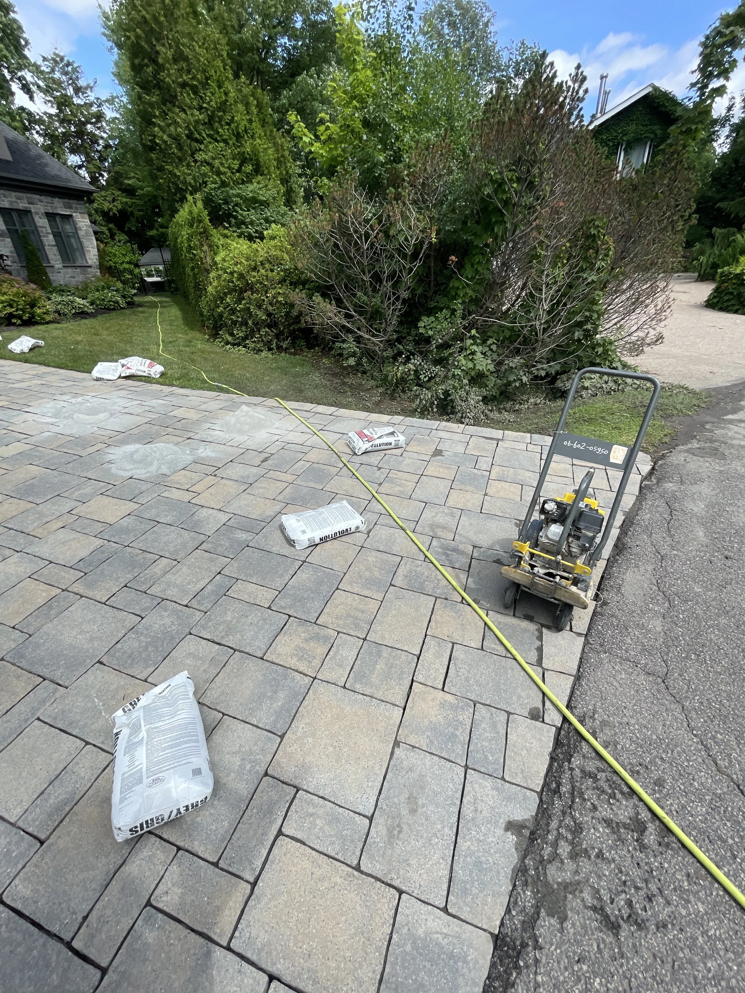 Travaux de pavage avec des sacs de matériaux et une machine à pavé sur une allée pavée, entourée de végétation et de maisons