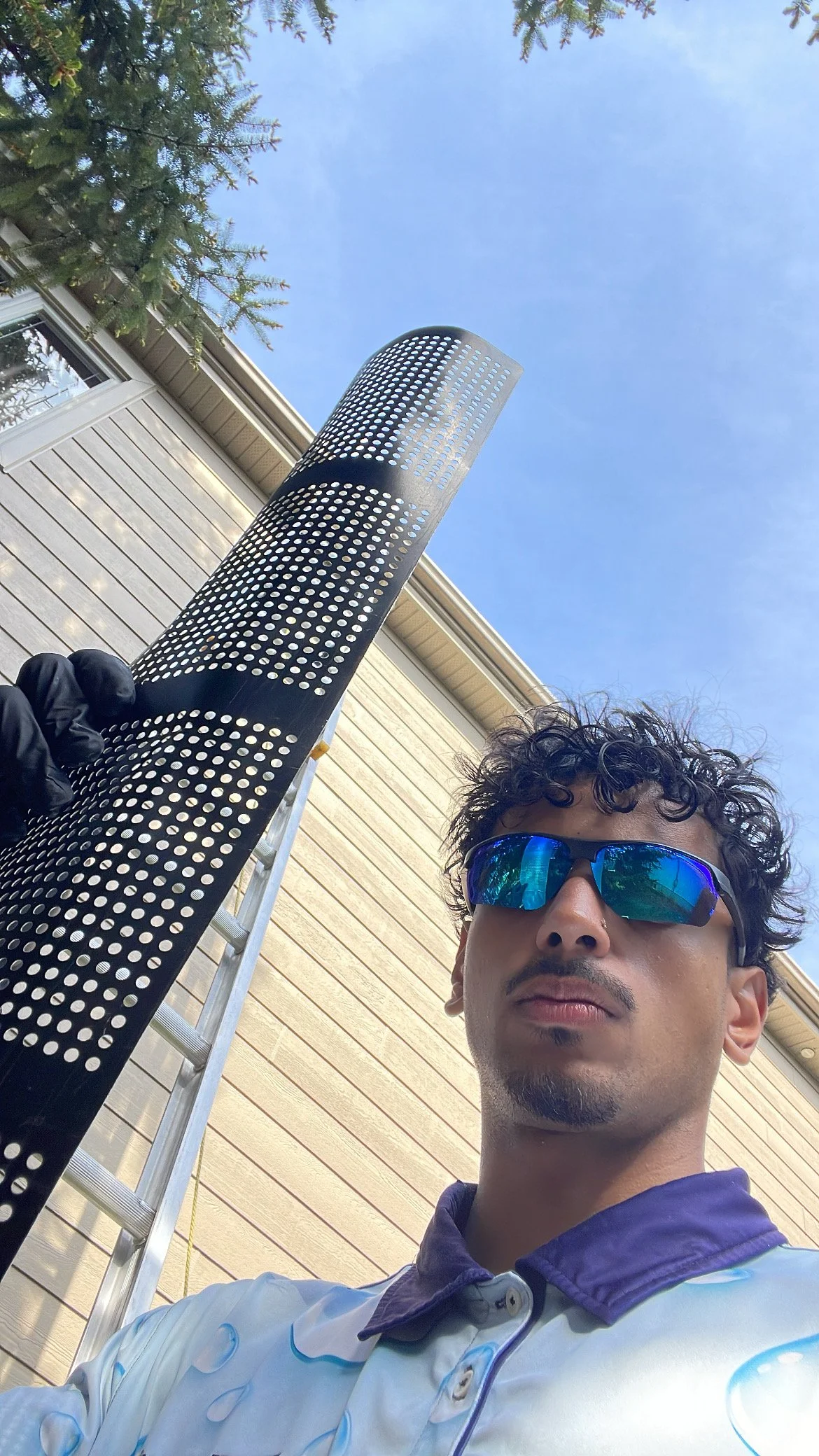 Un homme porte des lunettes de soleil réfléchissantes et une chemise à motifs. Il tient une rallonge perforée noire et se trouve à l'extérieur, près d'une maison en bois avec un toit en métal, sous un ciel bleu.