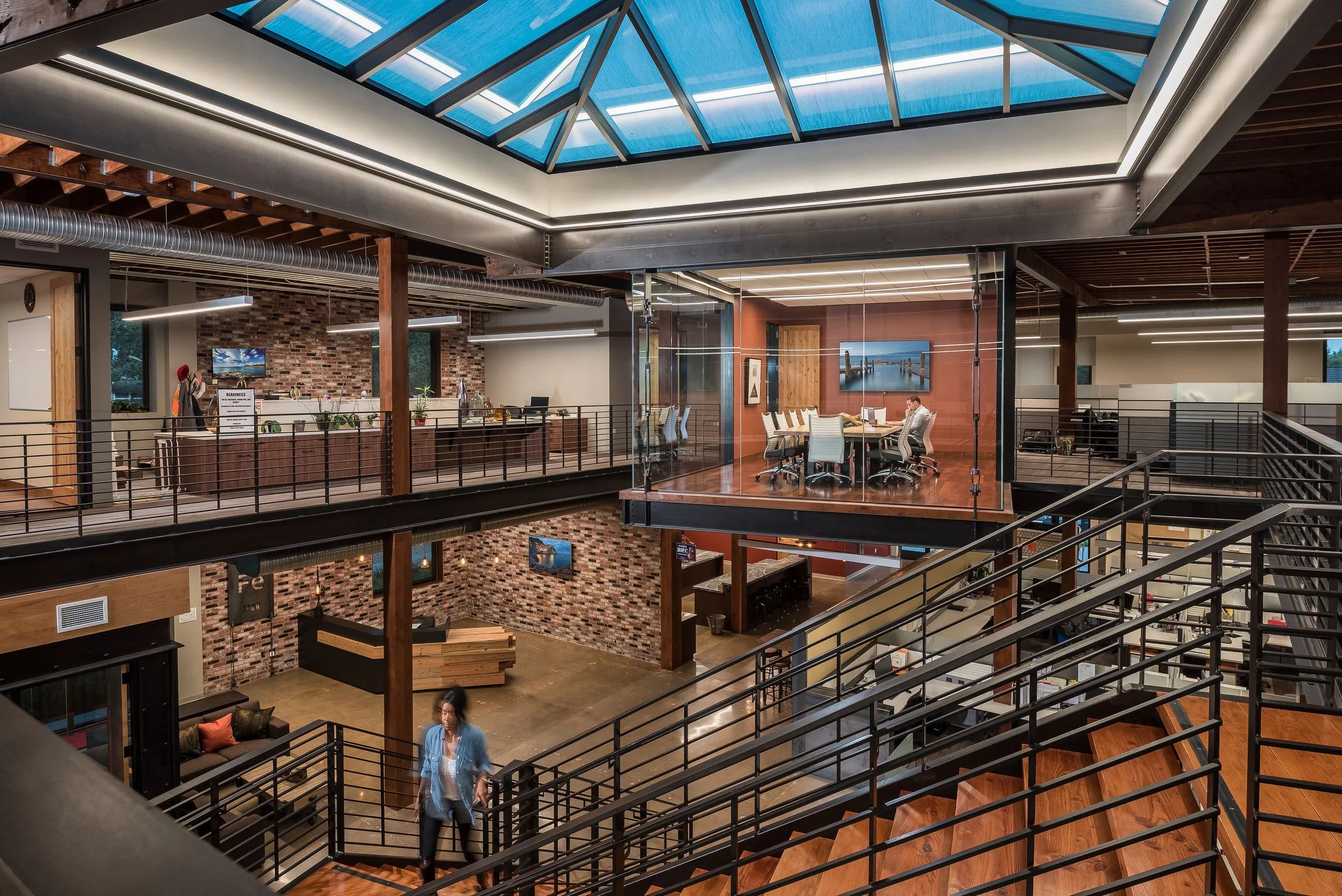 Interior of a modern, multi-story office with exposed brick walls, stairs, glass partitions, and a prowled conference room with a view of a dock and water.