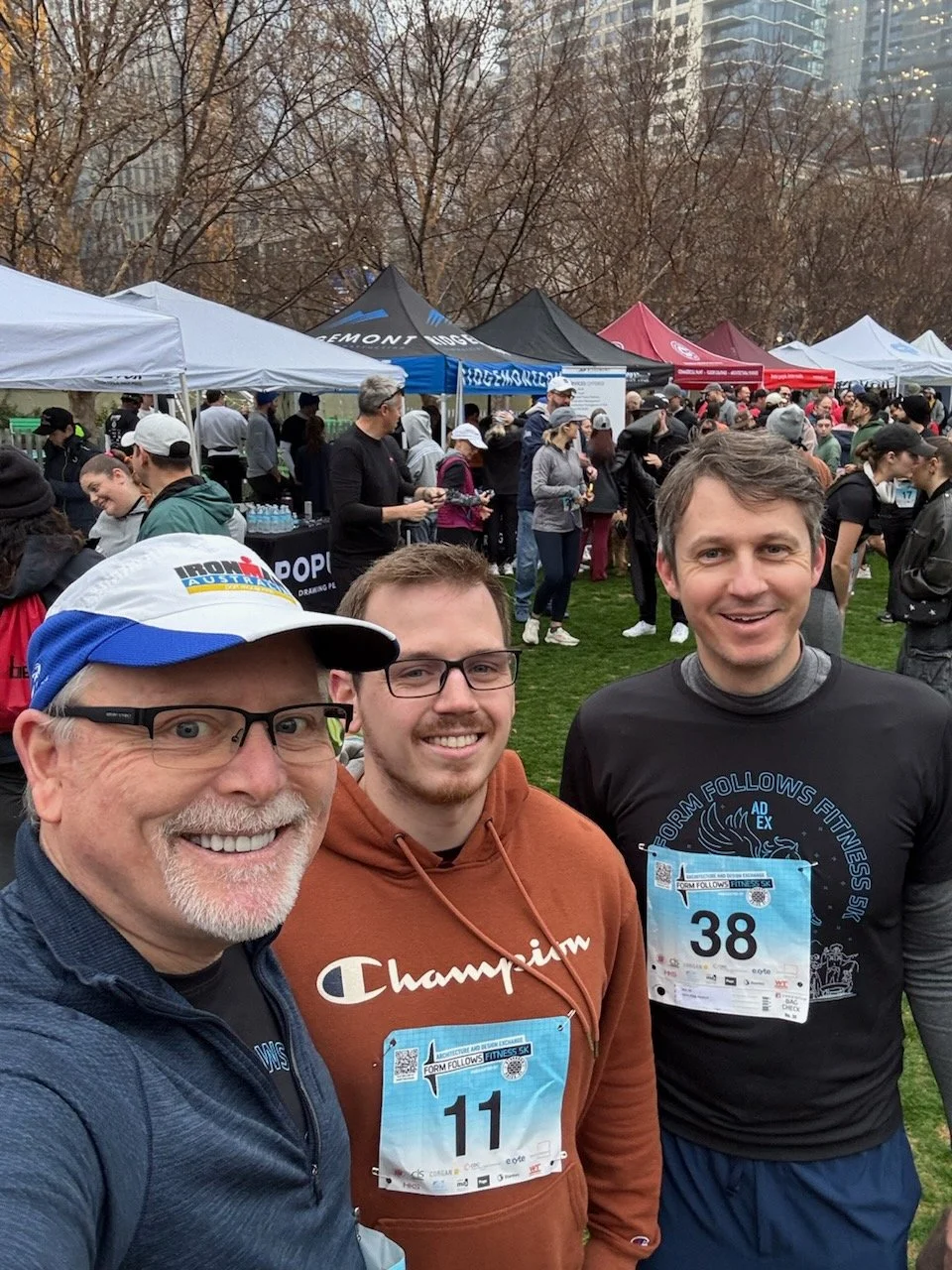 Three men smiling at a race event with bib numbers 11 and 38, surrounded by tents and people in athletic attire, trees in the background.