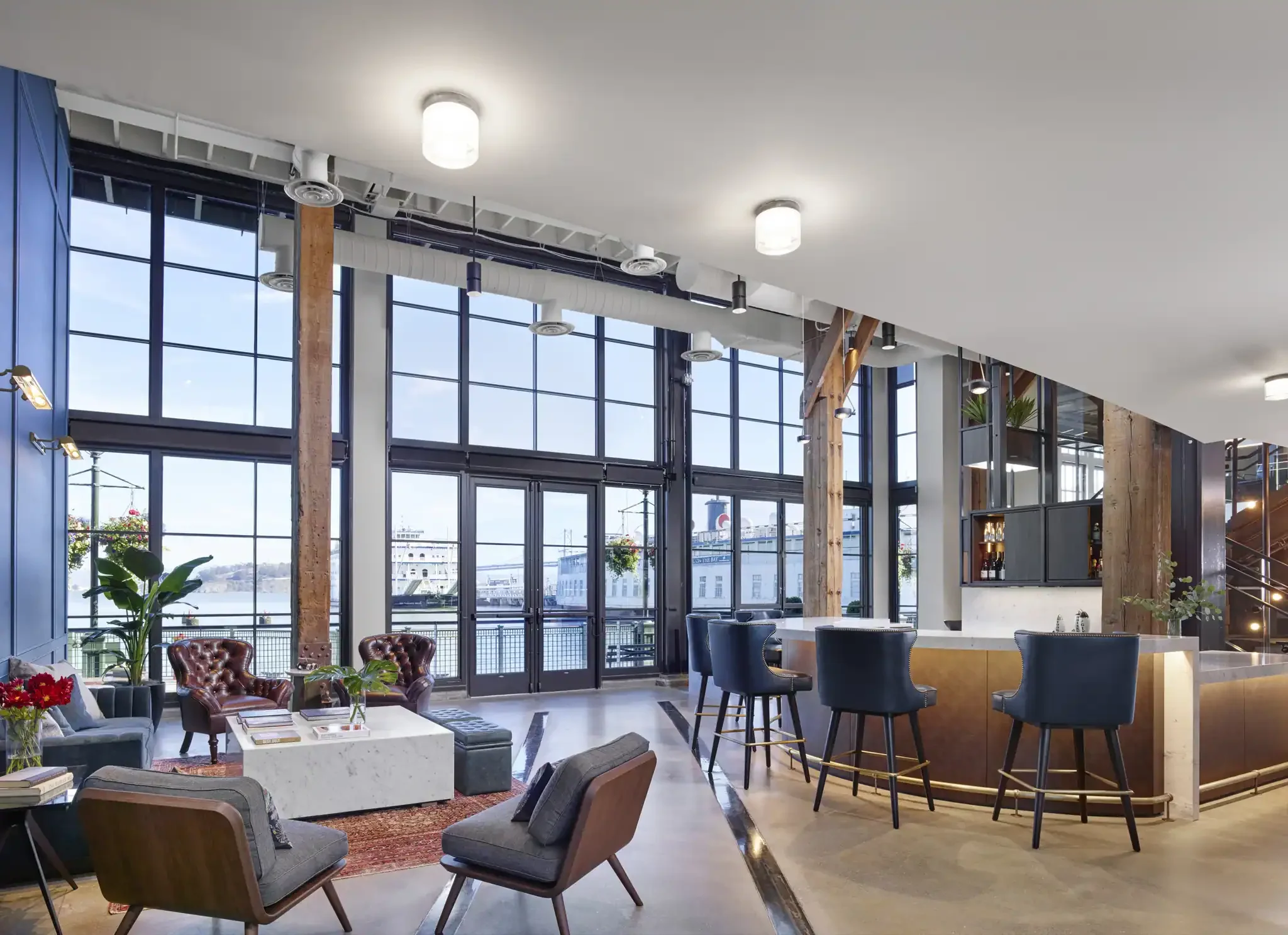 Interior view of the 8VC office lounge and bar area showcasing large industrial windows, wooden support columns, a bar with blue stools, and a seating area.