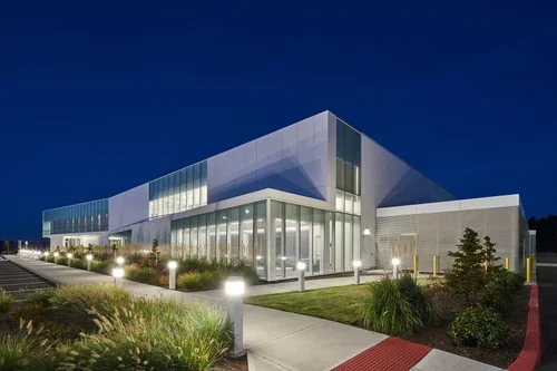 Modern building with large glass windows illuminated at night, surrounded by landscaped greenery and pathway lighting.